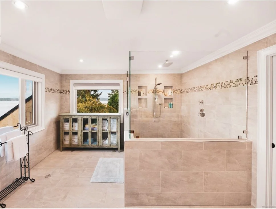Modern bathroom with beige tiled walls and floor, a glass-enclosed shower with built-in shelves, a window with a view of trees, and a white towel rack.
