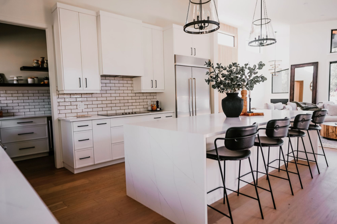Modern kitchen with white cabinets, a large island with black barstools, a black vase with greenery, and pendant lights.