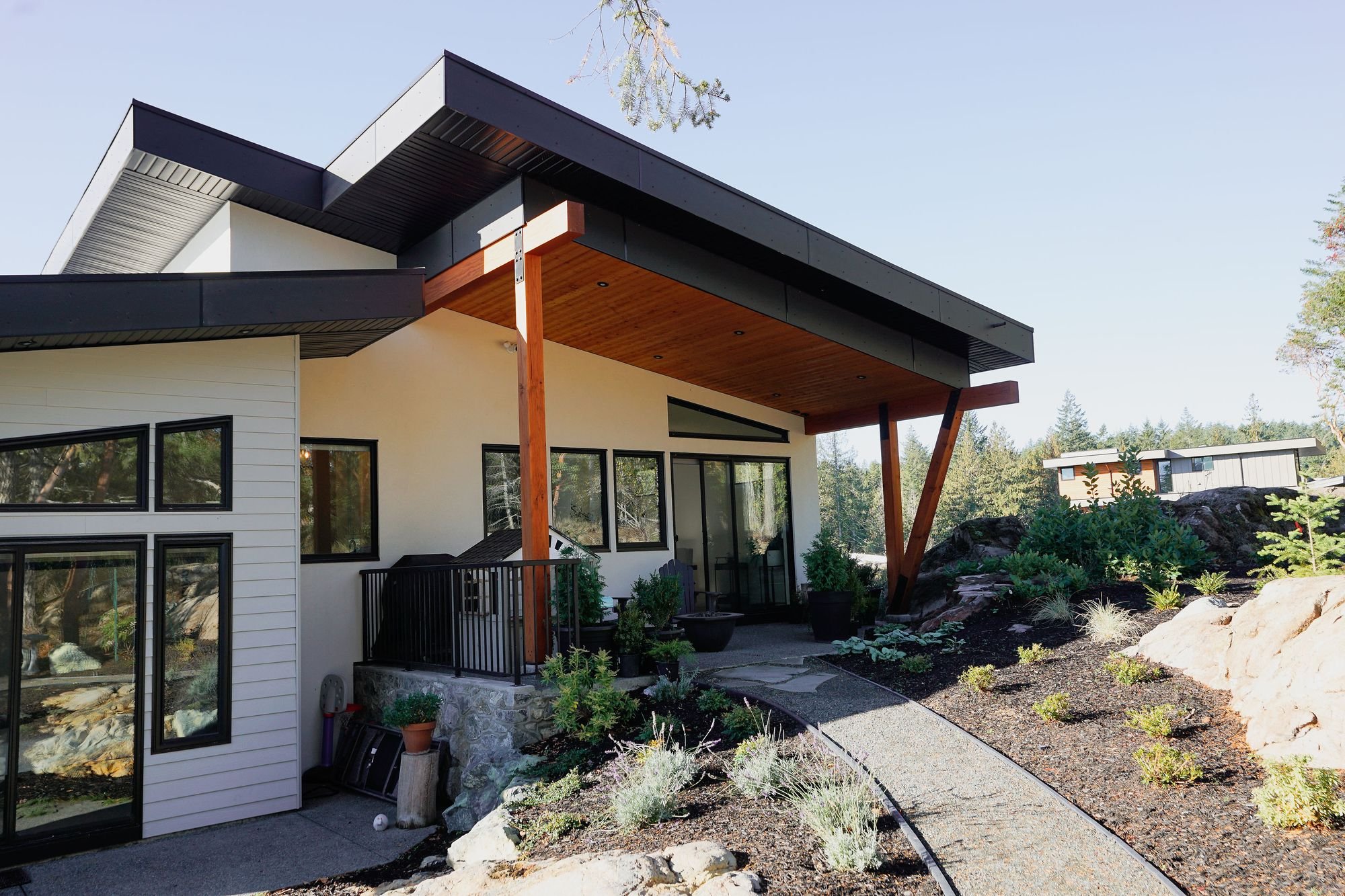 Modern house with black and white exterior, large windows, and a wooden balcony supported by angled wooden beams. The house has a landscaped yard with plants, rocks, and a pathway, surrounded by trees.
