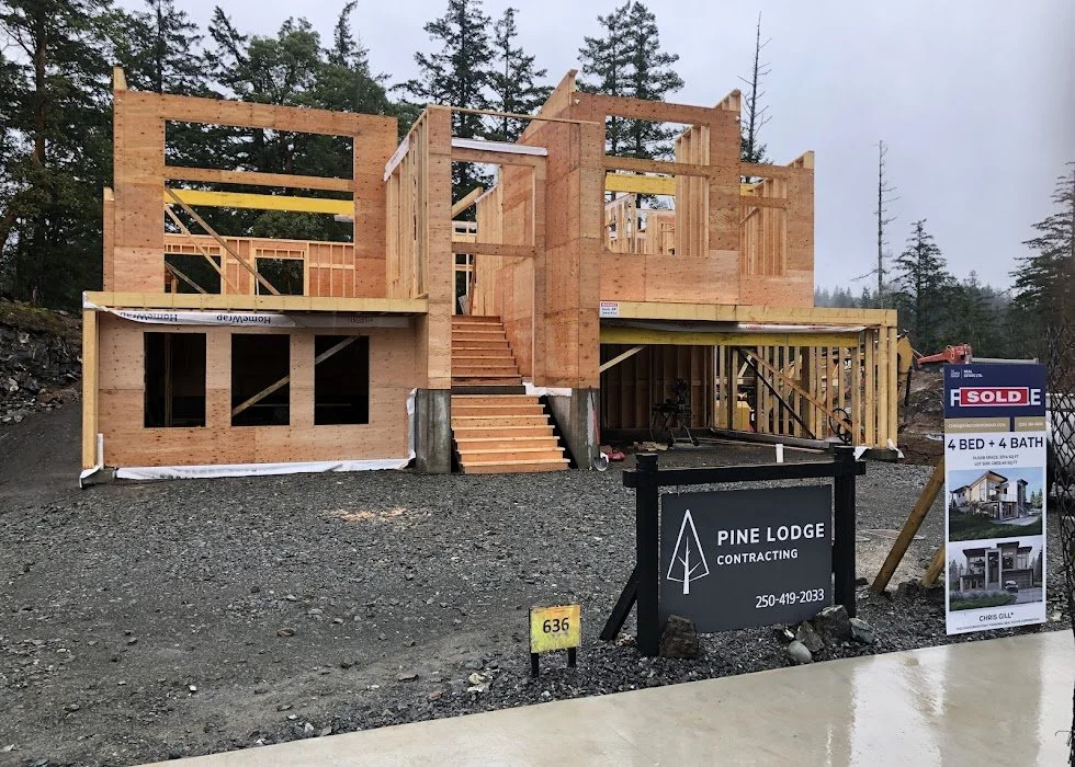 Under-construction two-story wooden house with framing and stairs, surrounded by trees, sign for Pine Lodge Contracting, and a 'sold' sign indicating a 4-bedroom, 4-bath home.
