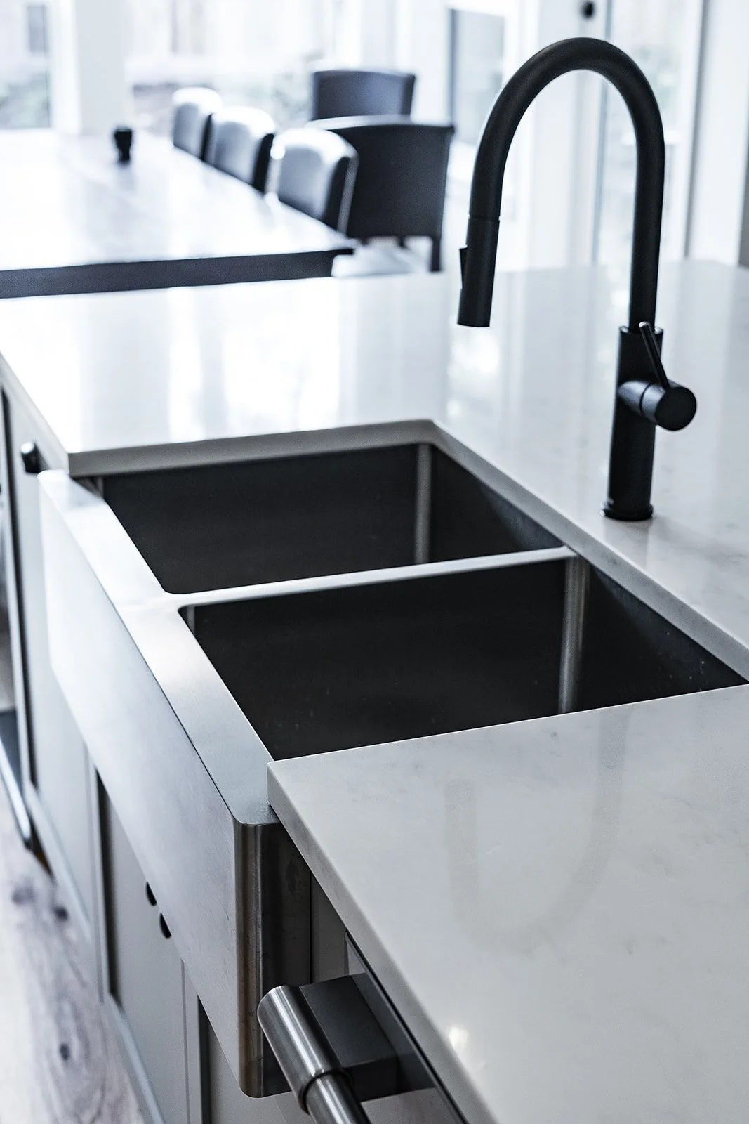 A modern kitchen sink with a black faucet on a white countertop, with a black under-sink compartment, in a bright kitchen with large windows.