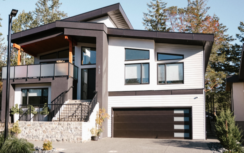 Modern two-story house with a black roof, white siding, and a black garage door. It has a front porch with steps and a balcony with a glass railing. Surrounding trees are visible in the background.