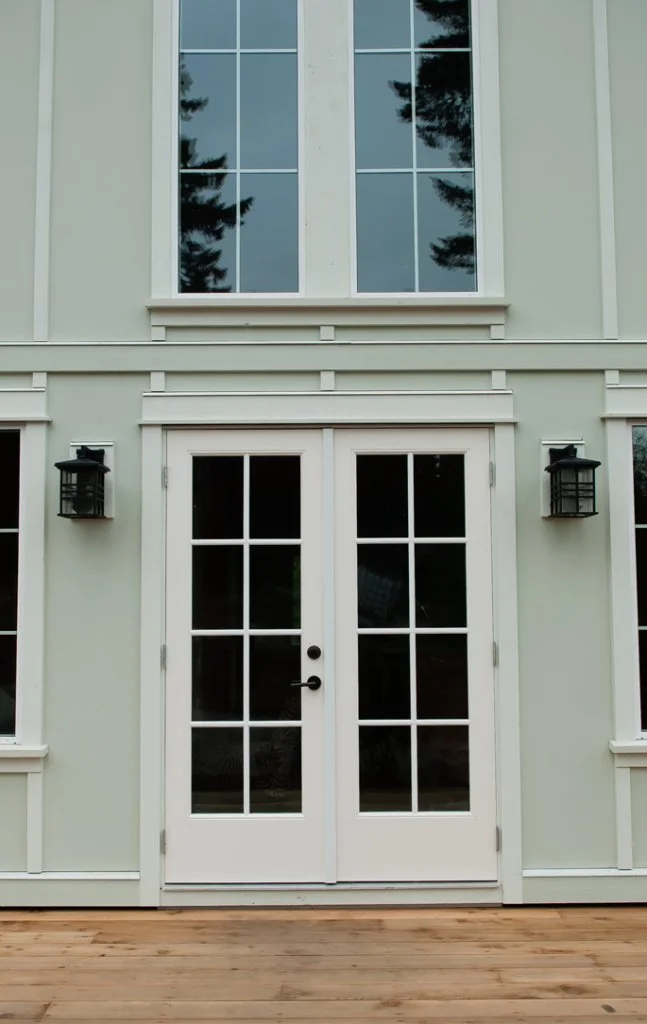 Exterior of a light-colored house with double French doors with glass panes, flanked by two black wall-mounted outdoor lanterns, and large upper windows reflecting trees, with a wooden deck in the foreground.