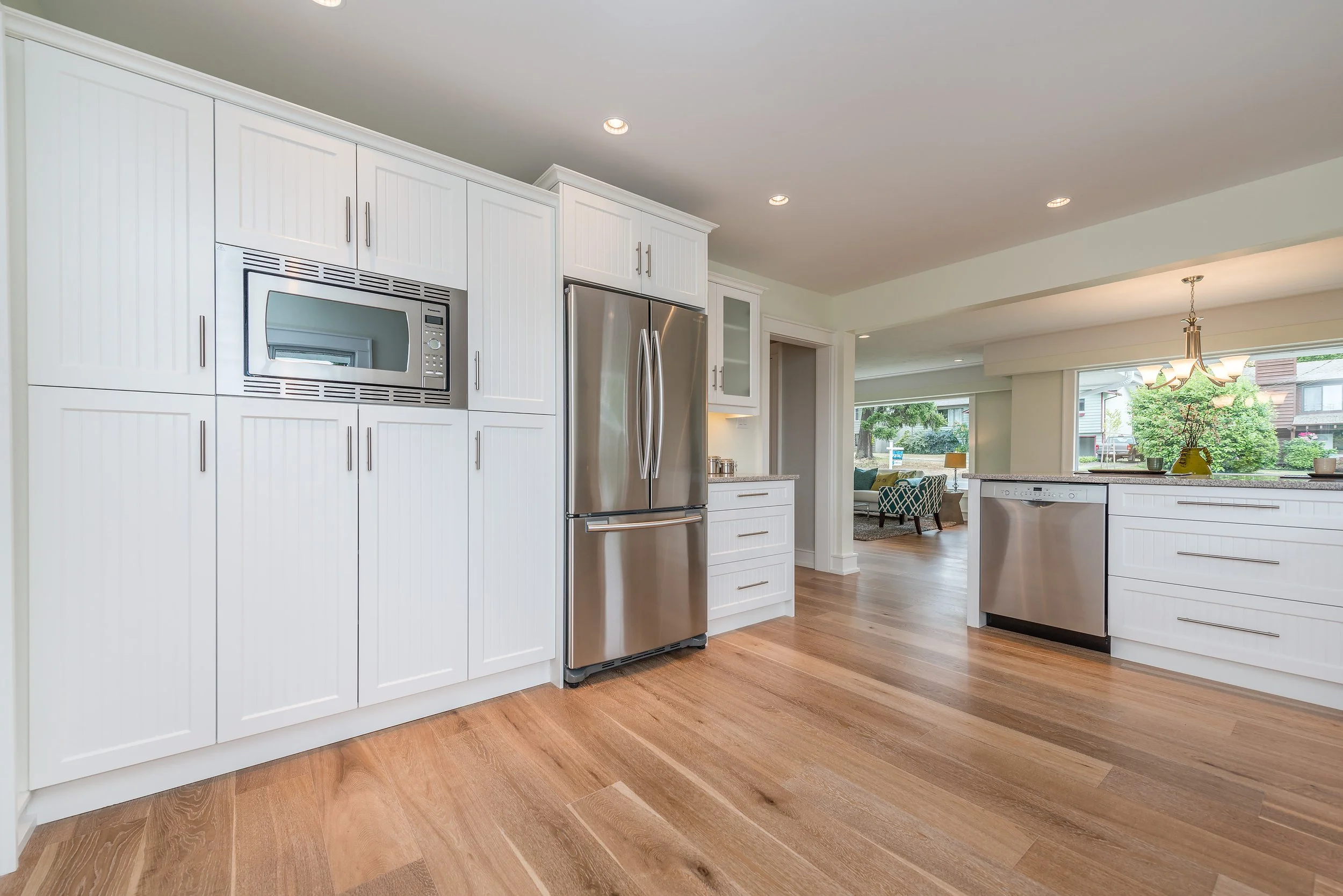 Modern kitchen with white cabinets, stainless steel appliances including a refrigerator, microwave, and dishwasher, hardwood floors, and large windows opening to a backyard.