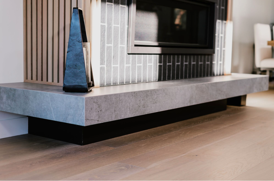 Modern living room with a concrete fireplace mantel, black LED TV, and decorative items on the mantel. Hardwood flooring and a partial view of a dining area in the background.