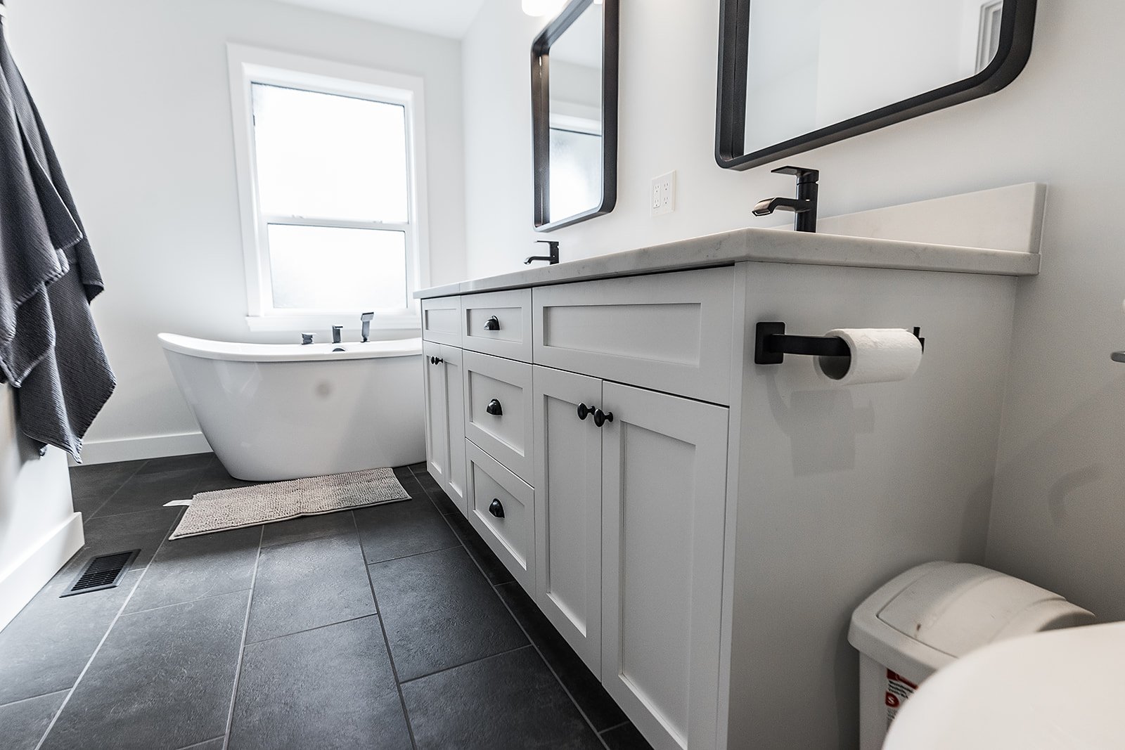 Modern white bathroom with a dark gray tile floor, a white vanity with black hardware and two mirrors, a bathtub under a window, and a toilet with a toilet paper roll holder.