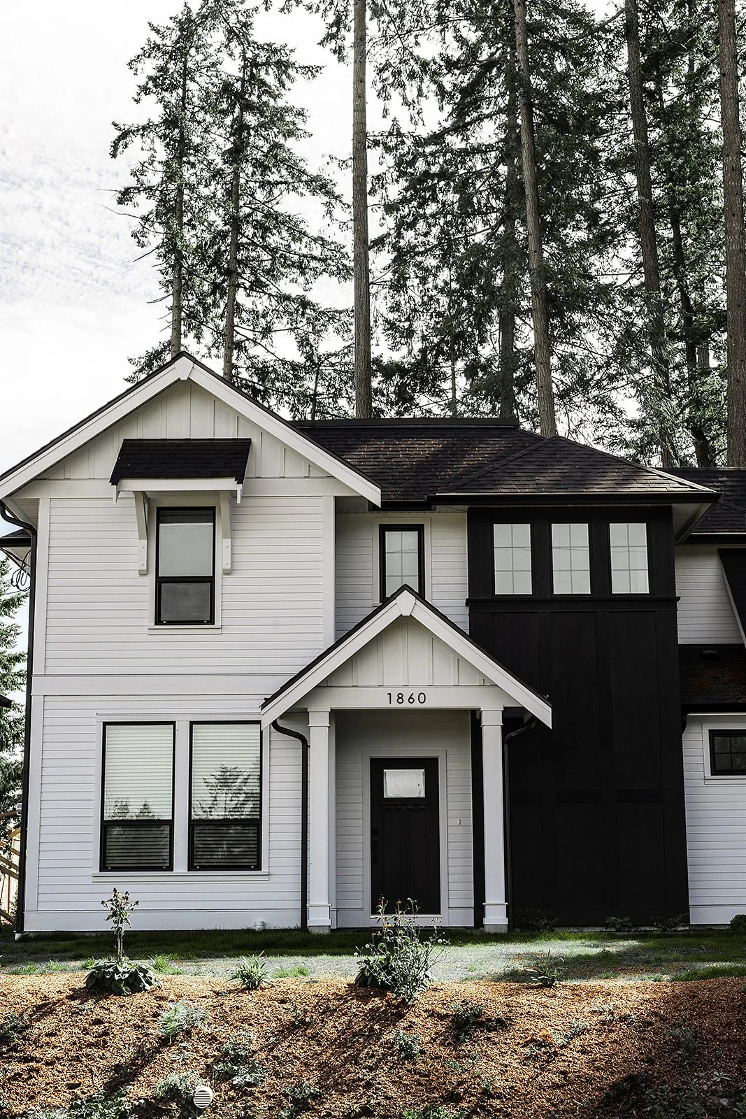 Front view of a two-story house with white siding, black accents, and a gabled roof, surrounded by tall trees.