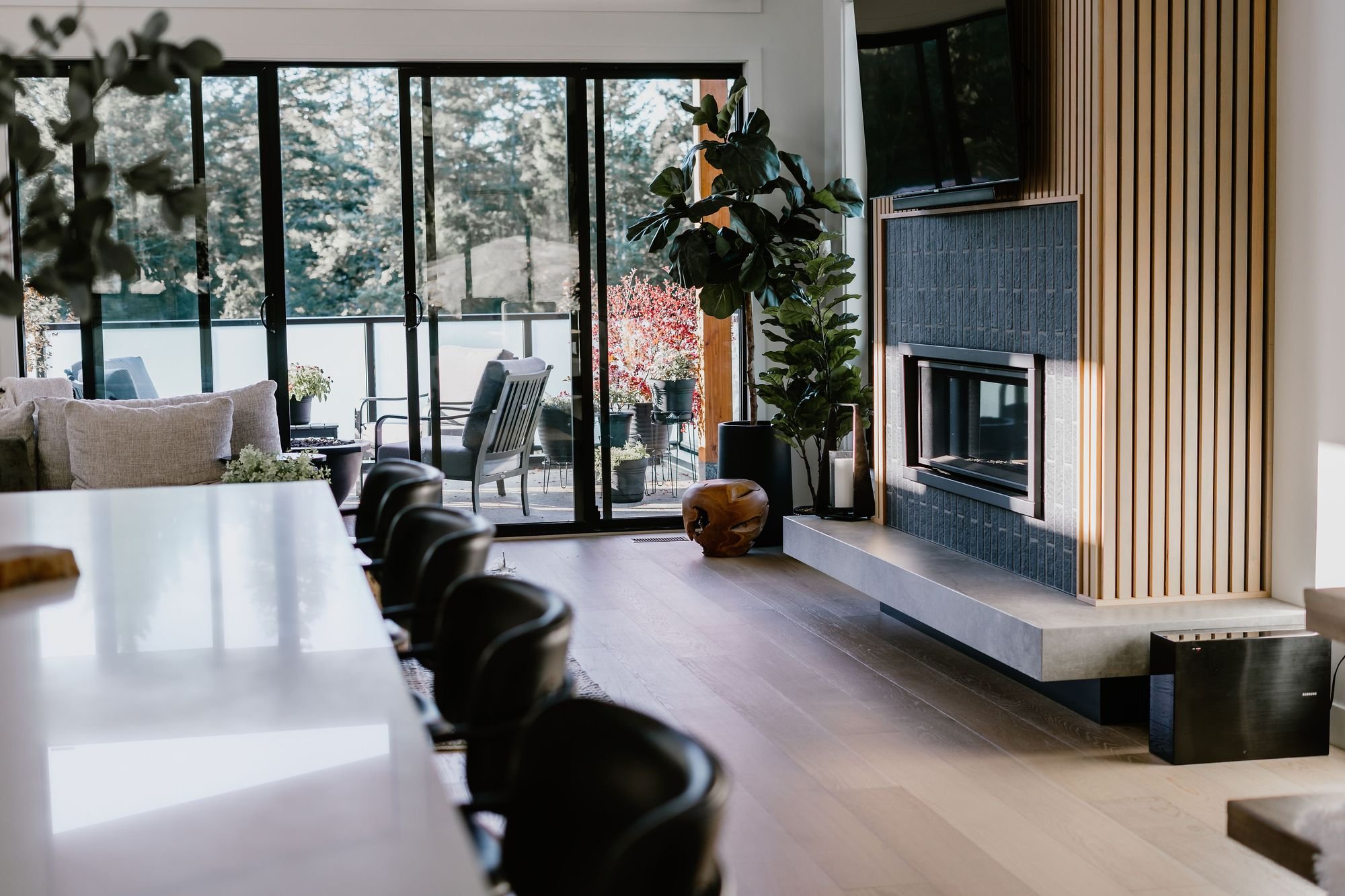 Modern living room with floor-to-ceiling sliding glass doors opening to a balcony with outdoor chairs and potted plants, a cozy fireplace with wooden slats on the wall, and a large plant next to the fireplace.