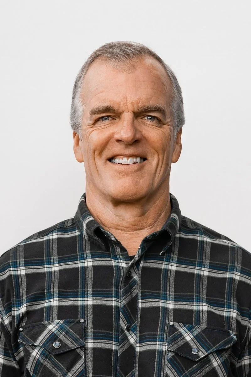 A middle-aged man with short gray hair, blue eyes, and light skin, smiling and wearing a black, gray, and blue plaid button-up shirt against a plain white background.