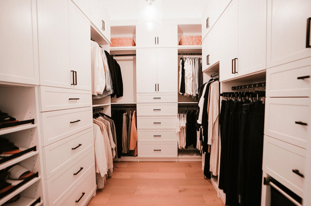 Walk-in closet with white built-in shelves filled with clothing, shoes, and pink storage boxes, hardwood floor, and well-lit space.
