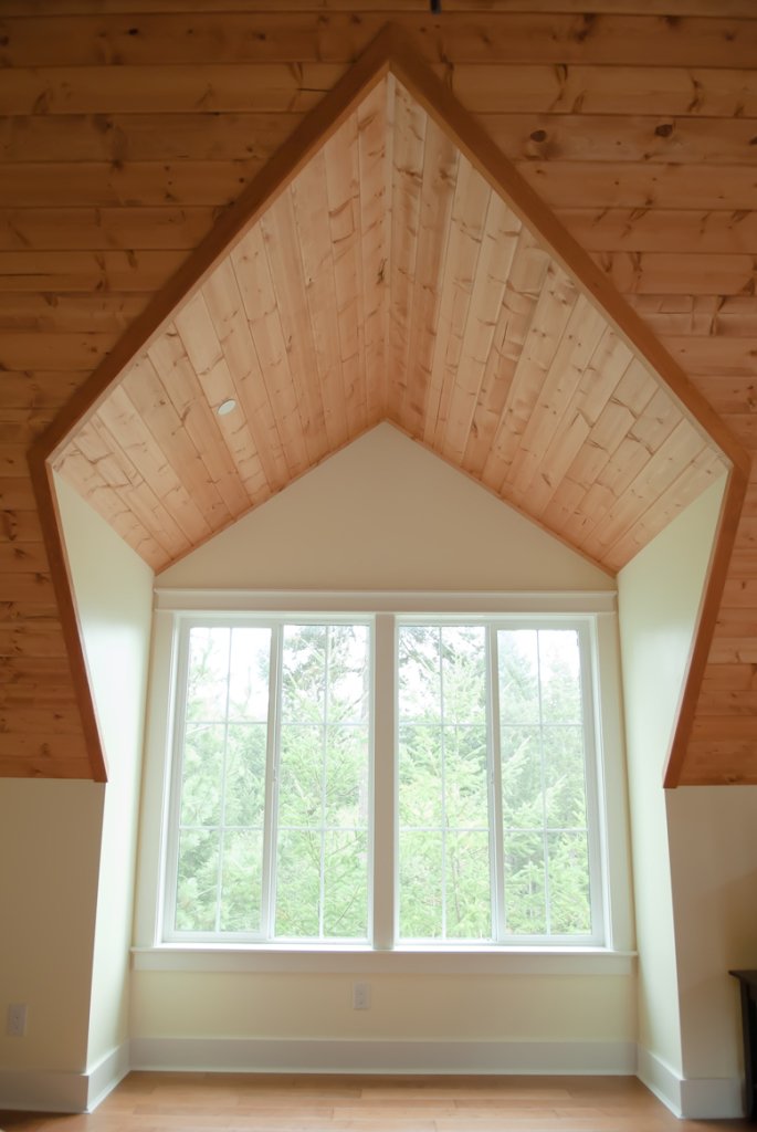 Interior view of a room with a vaulted ceiling covered in wood paneling, large window with multiple panes, overlooking green trees outside.