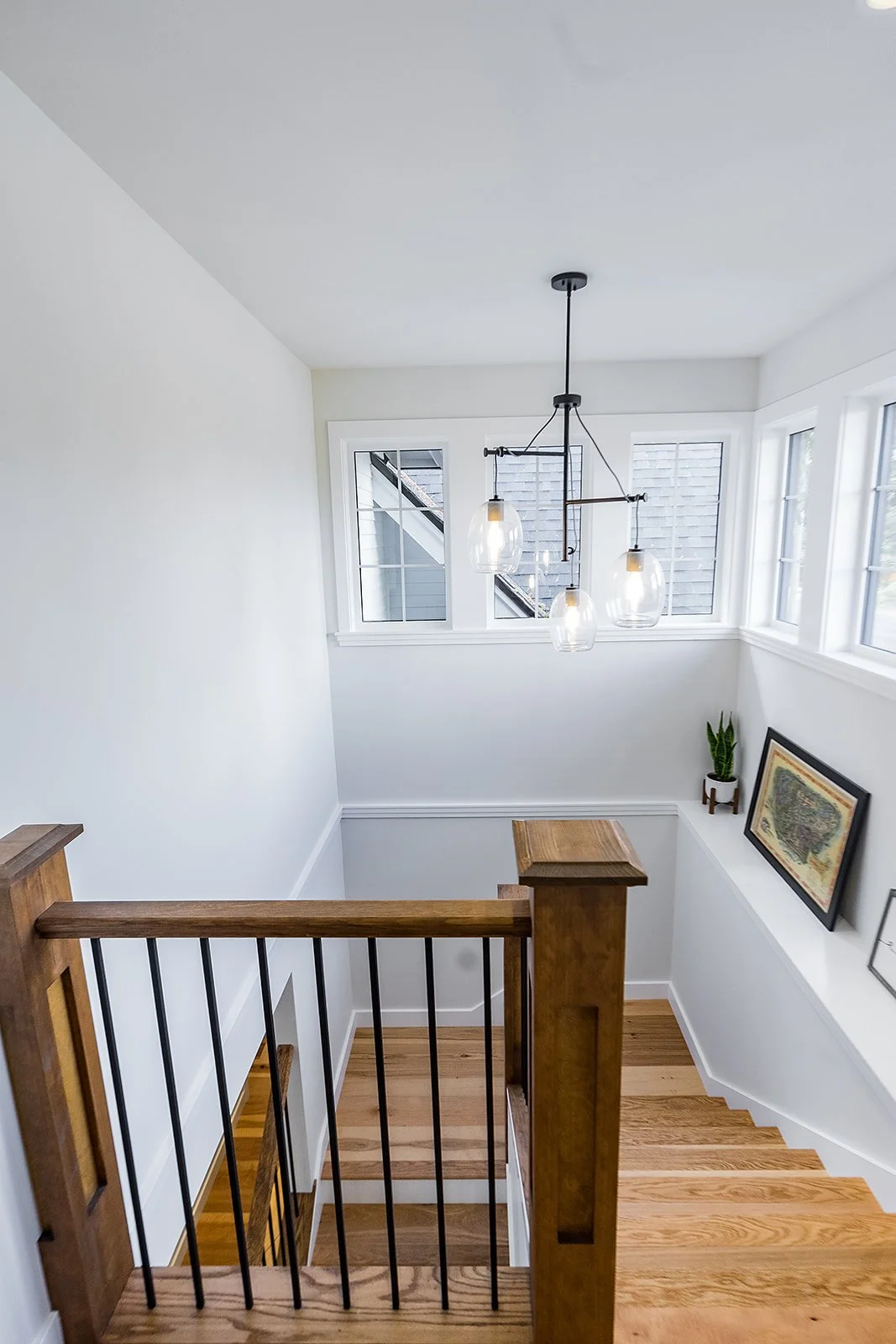 View of a staircase landing with wooden steps, white walls, a modern chandelier with three glass pendant lights, three large windows, a framed map, and a potted plant.