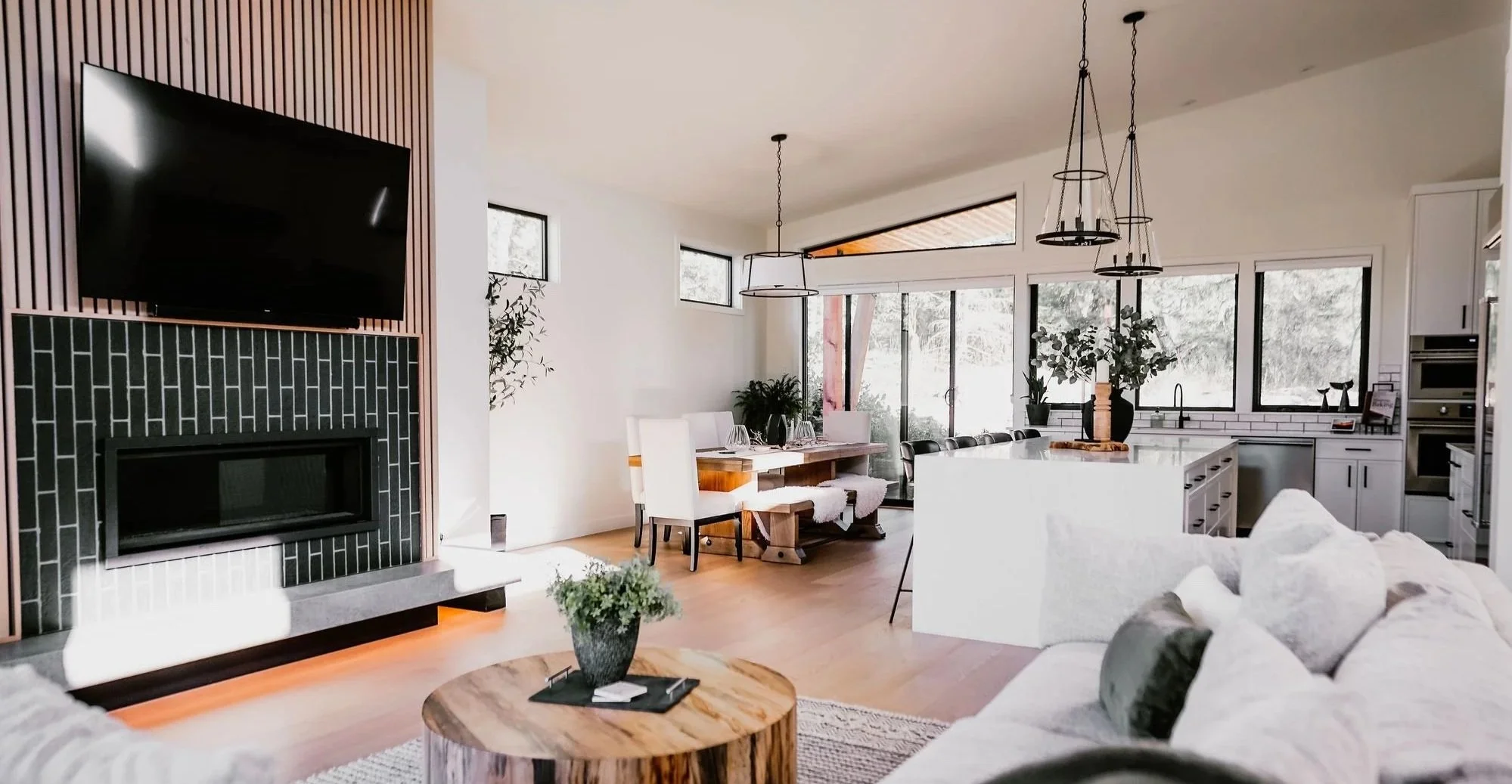 Open-plan living space with a mounted TV above a black tiled fireplace, a dining area with white chairs and a wooden table, and a kitchen with large windows, white cabinets, and modern appliances. Decor includes plants and pendant lighting.