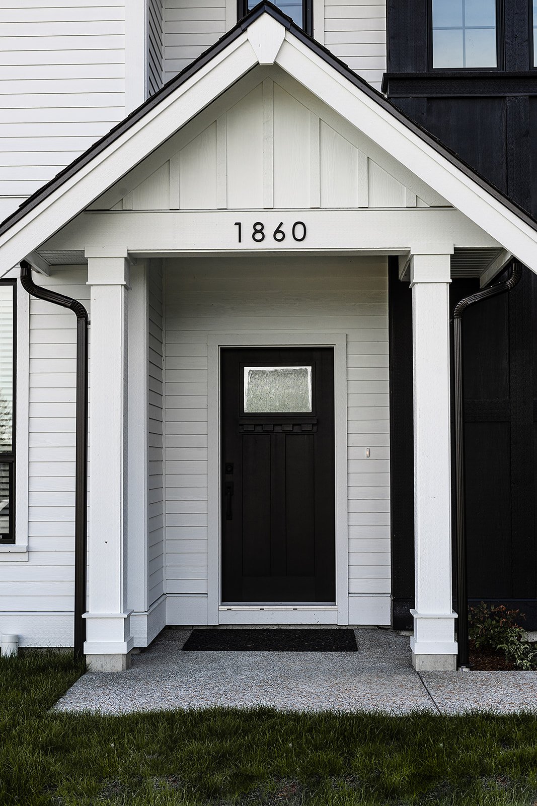 Close-up of a modern house entrance with the year 1860 displayed above the door, featuring white and black siding, a black door with a window, and a small porch with a concrete walkway.