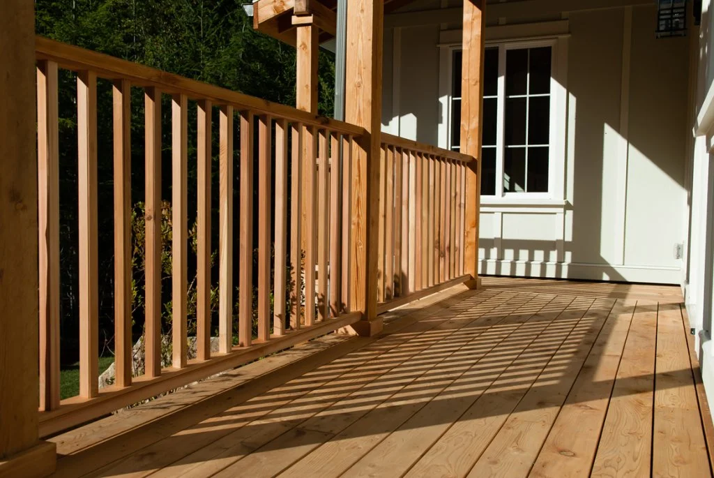 Wooden porch with railing and a white door with a large window, sunlight casting shadows on the floorboards.