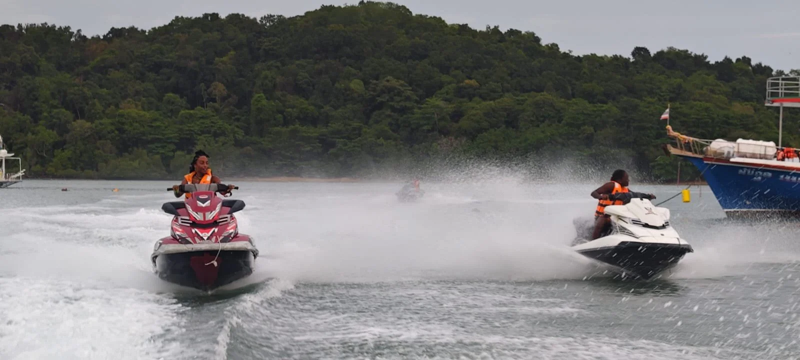 Two people riding jet skis on a body of water near a wooded shoreline, with boats in the background.
