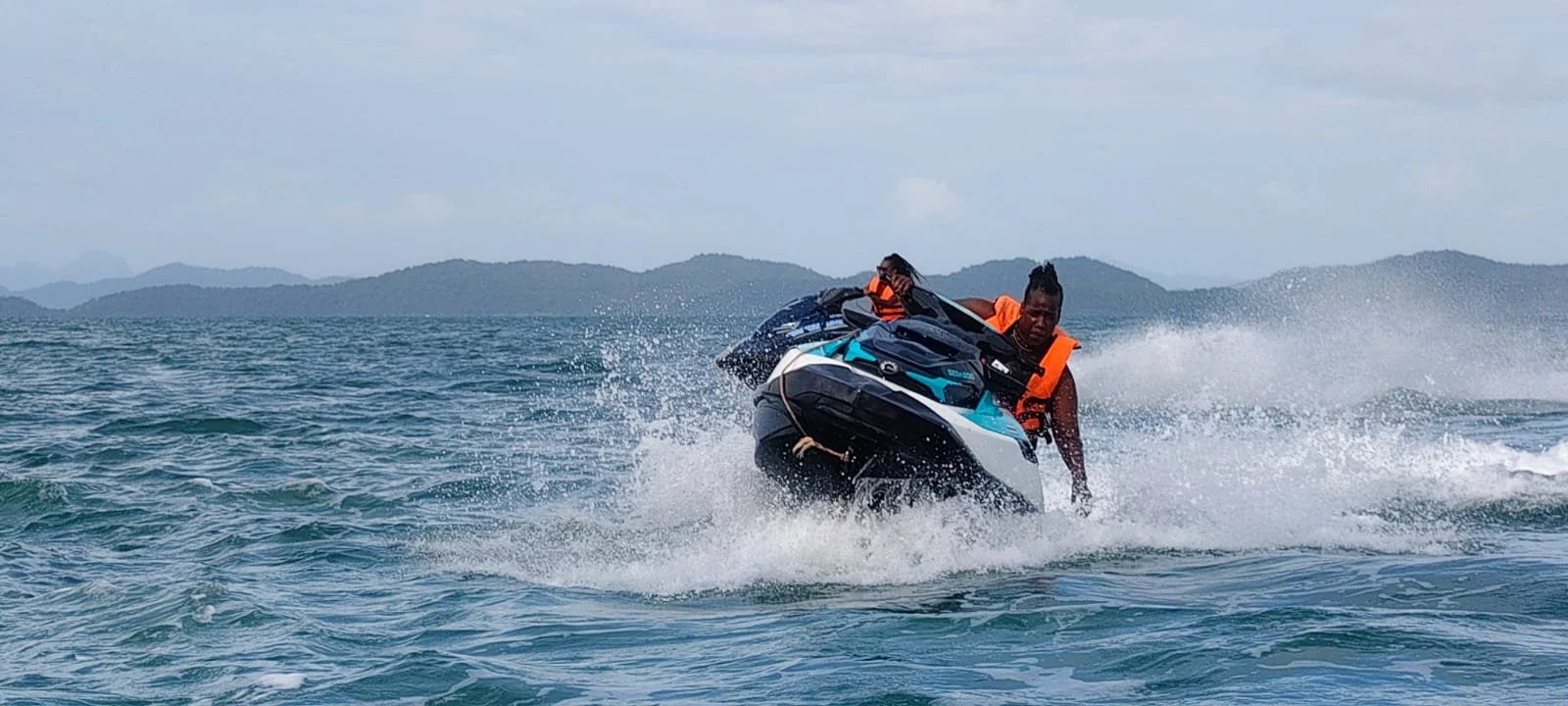 Two people riding a jet ski on the water with an island or mountain range in the background, wearing orange life jackets.