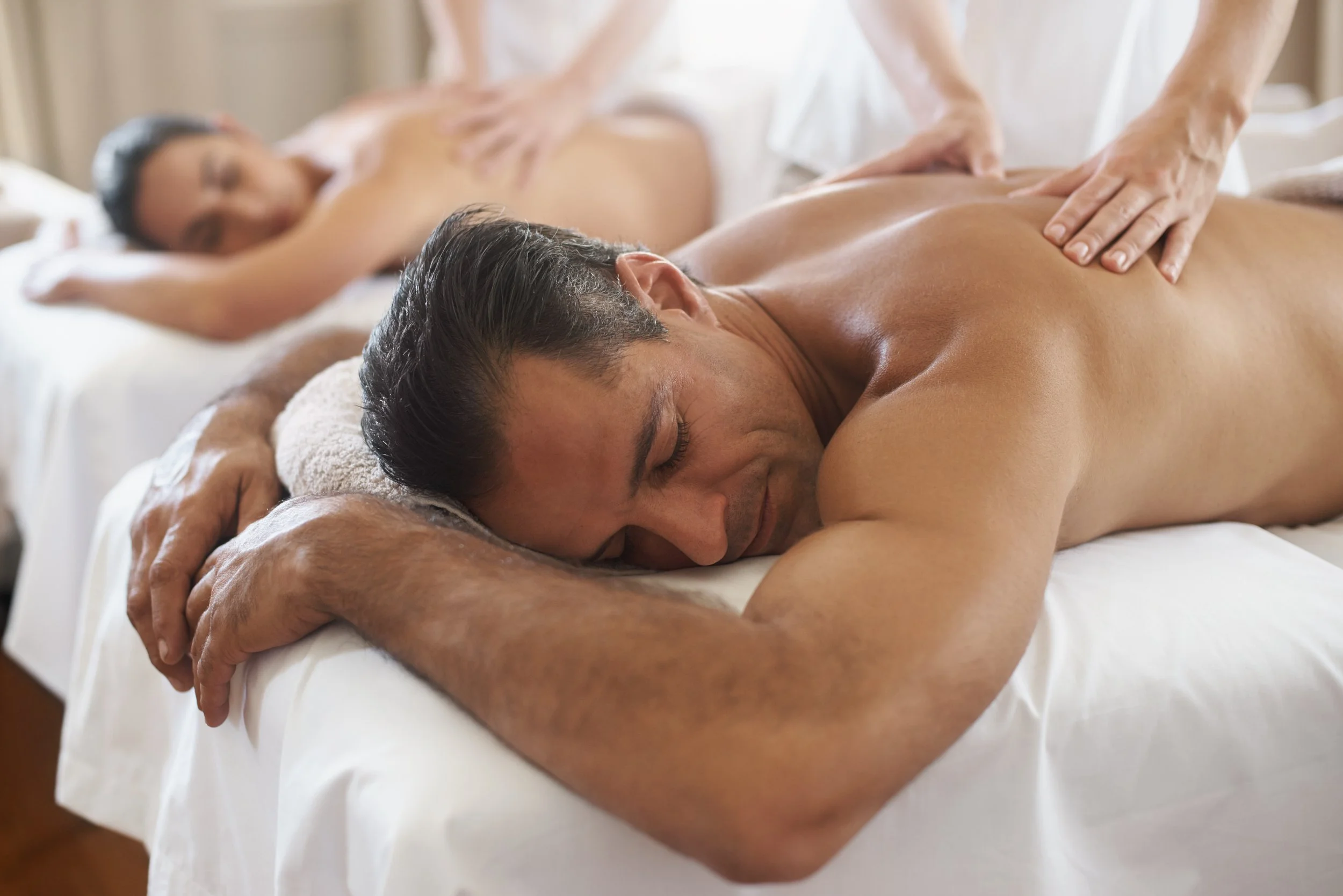 Two people lying face down on massage tables receiving massages from therapists in a well-lit room.