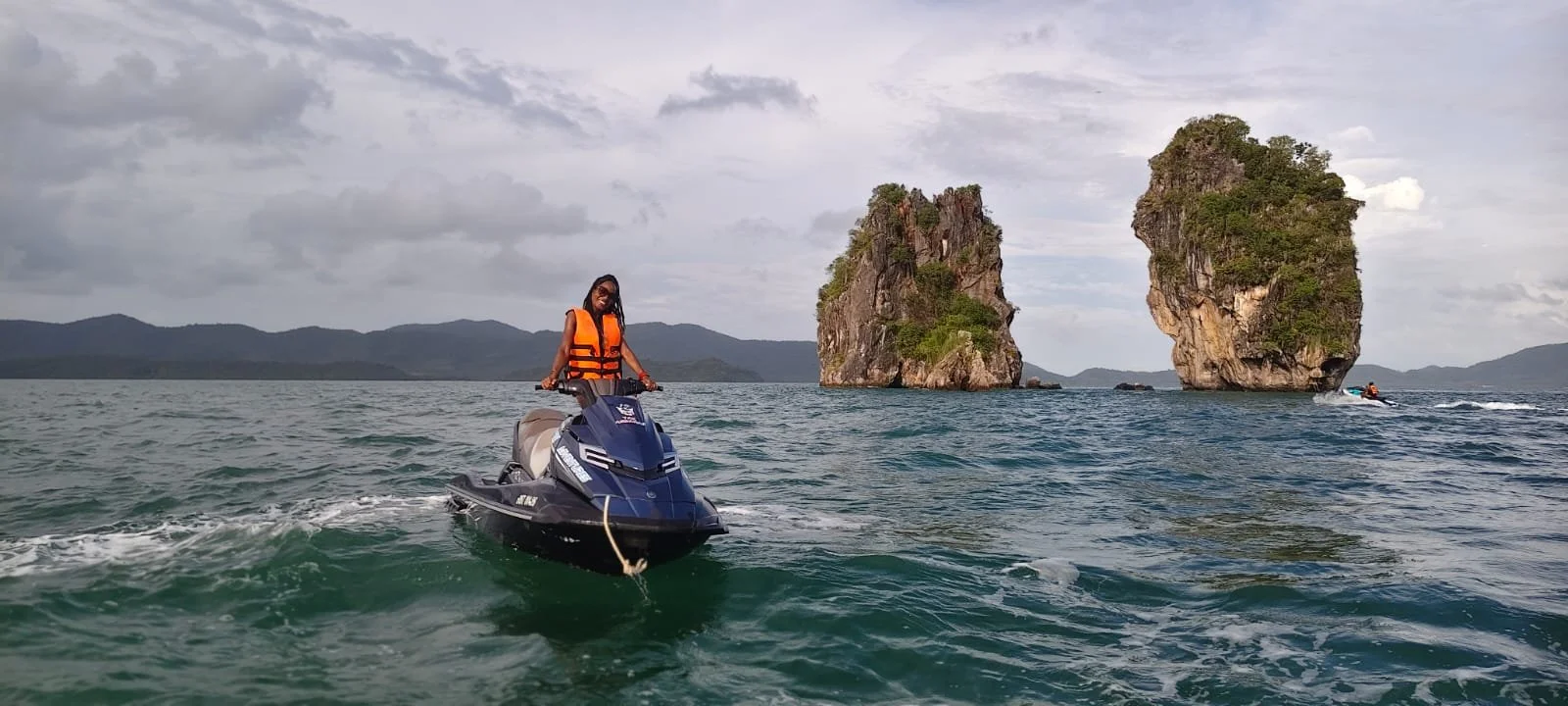 A woman in an orange life jacket rides a jet ski on the water with rocky islands and distant mountains in the background.