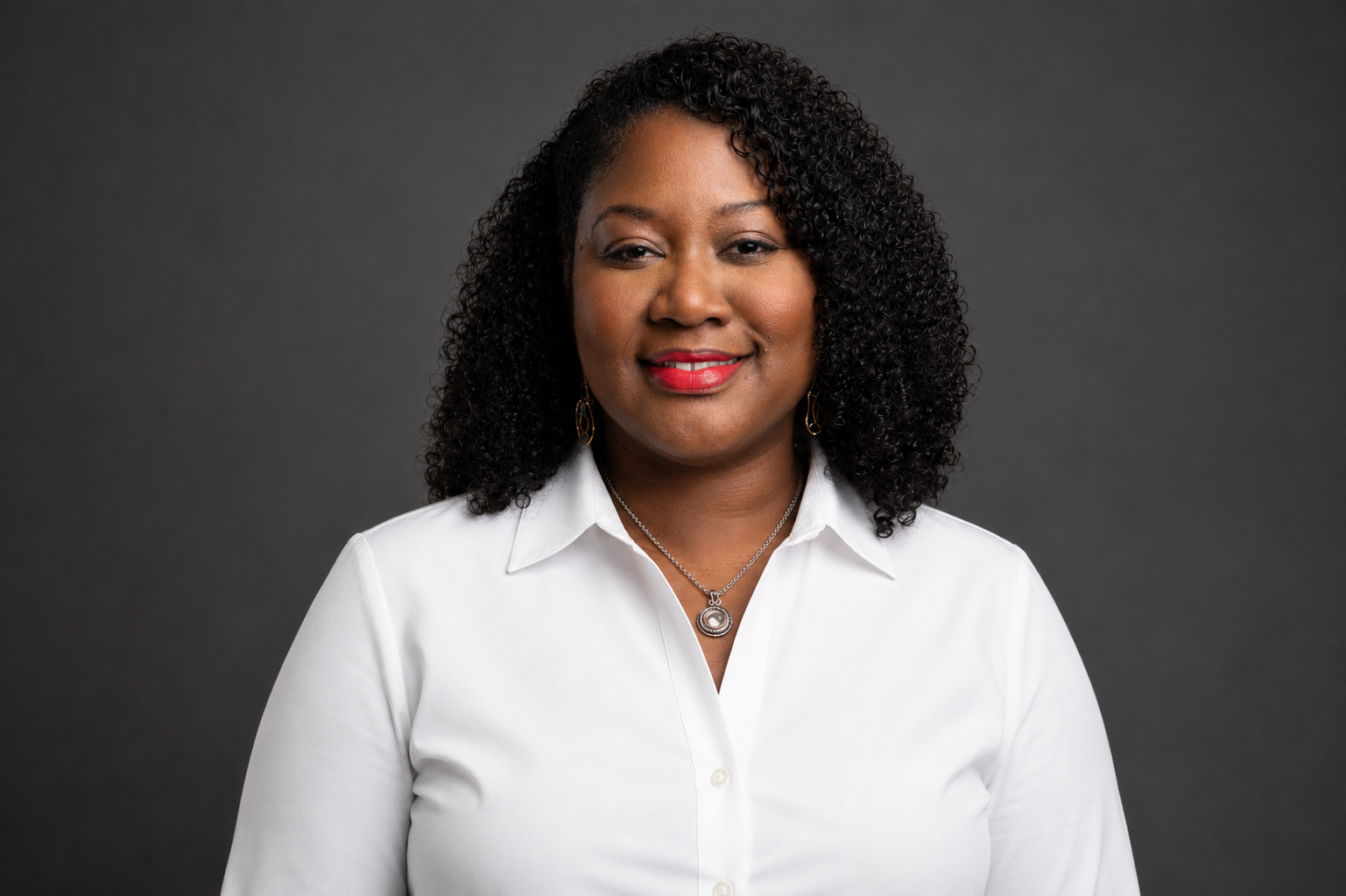 Portrait of a woman with curly black hair wearing a white shirt, red lipstick, earrings, and a necklace, smiling against a dark gray background.
