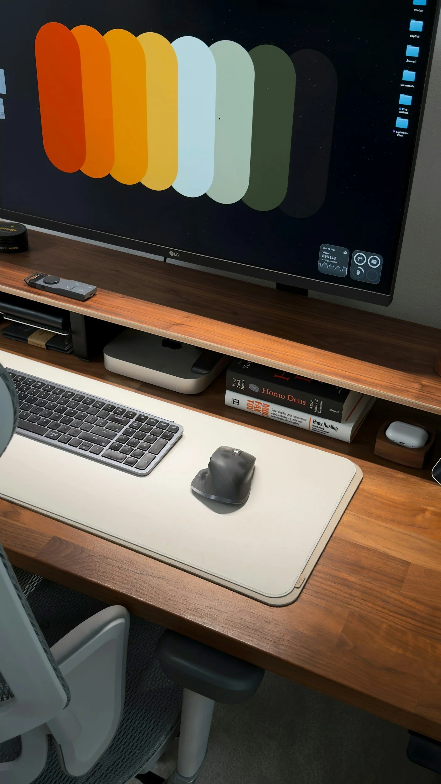 A computer desk setup shows a large monitor displaying a colorful abstract wallpaper, a wireless keyboard, a vertical mouse, a computer mouse pad, a small computer, a few books, a remote, and a white chair with a black mesh seat.