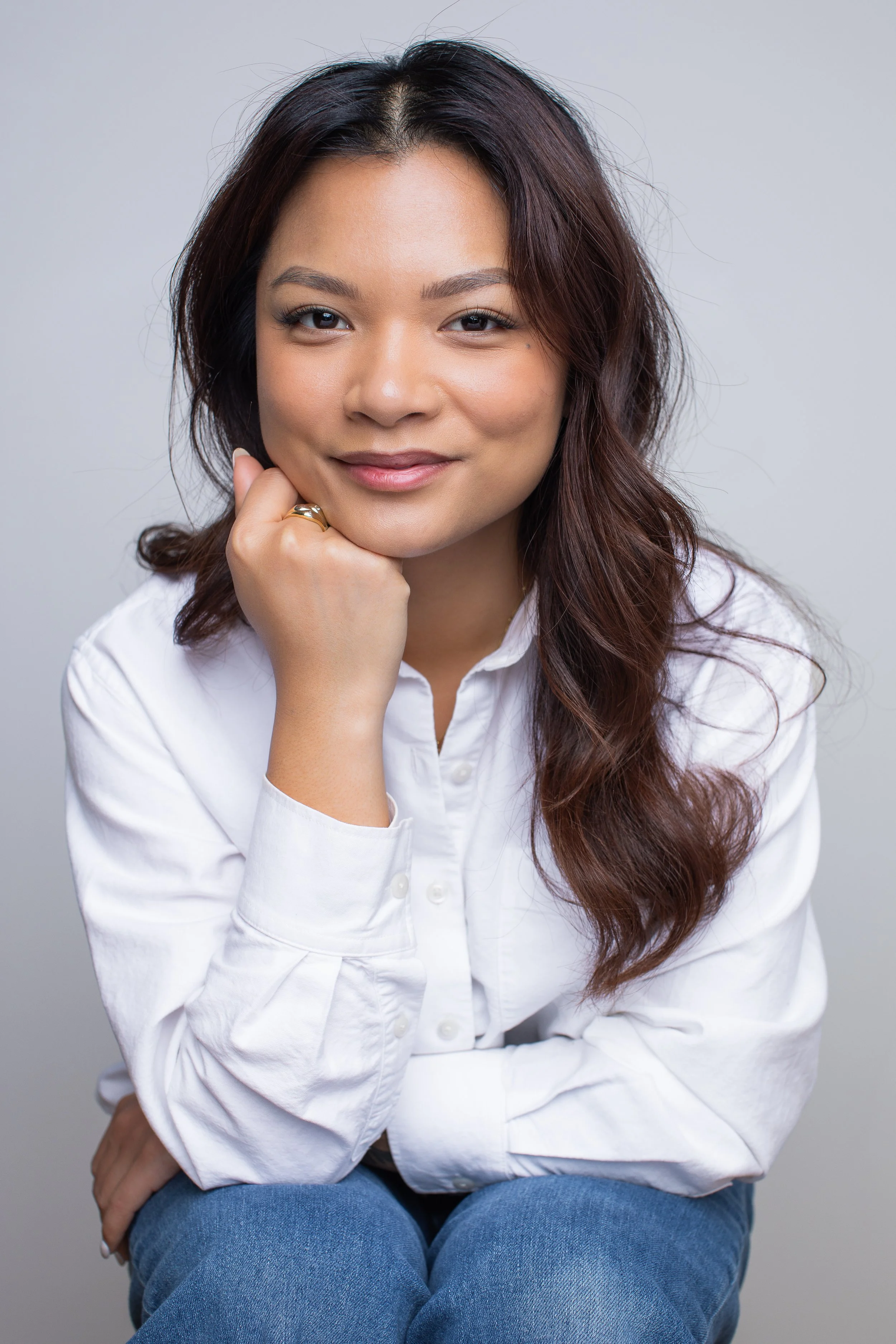 A woman with long wavy brown hair, wearing a white button-up shirt and blue jeans, sits with her chin resting on her hand and smiles at the camera against a plain gray background.