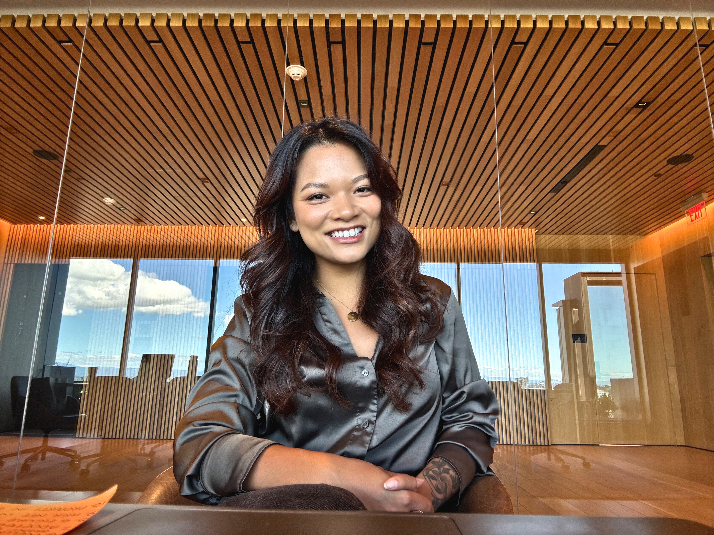 Portrait of a young woman with long dark wavy hair and a smile, sitting indoors with a wooden ceiling and large windows in the background.