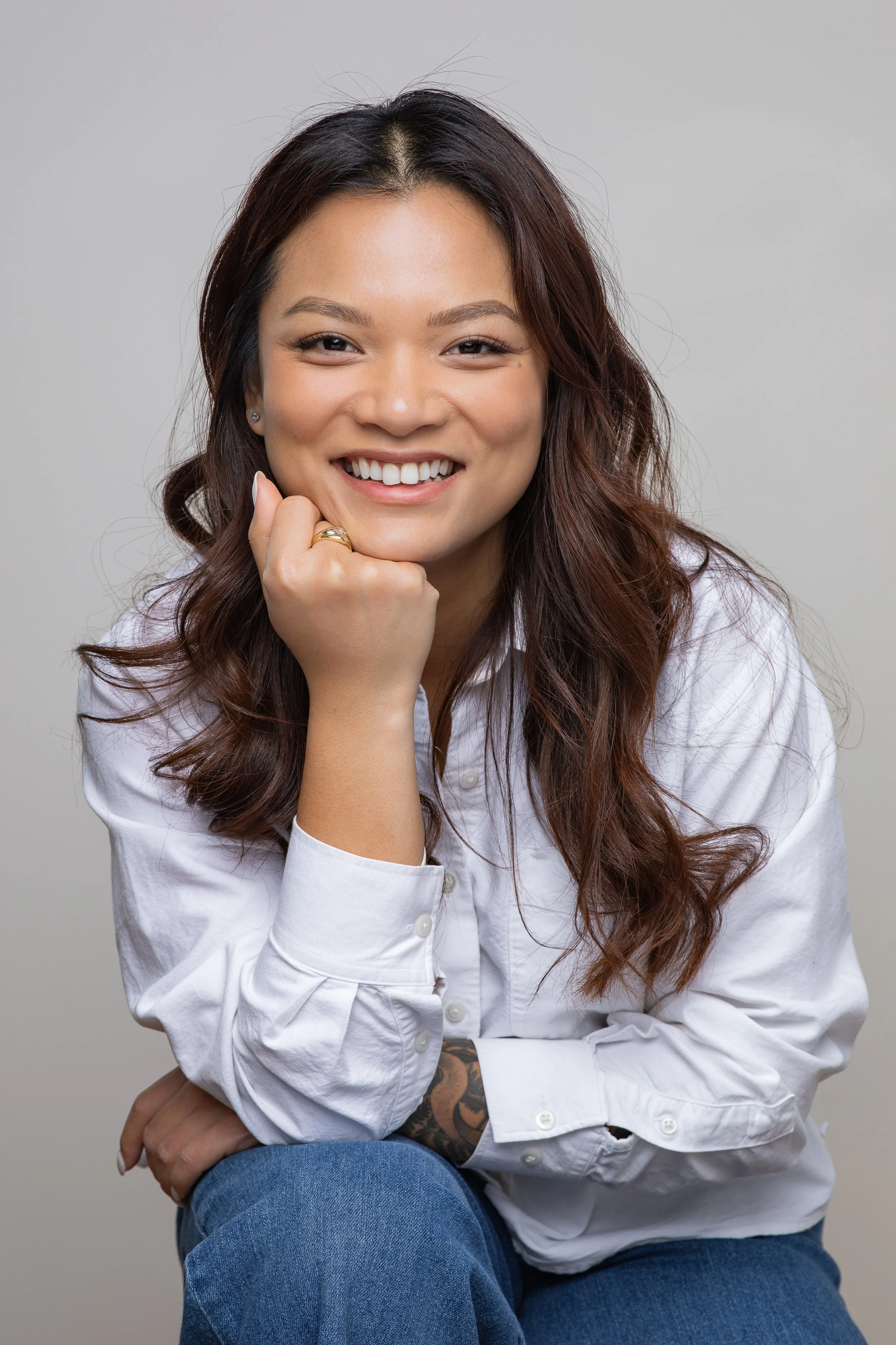 Portrait of a woman with long brown hair, smiling, wearing a white button-up shirt and blue jeans, sitting with her hand resting on her chin, against a plain gray background.