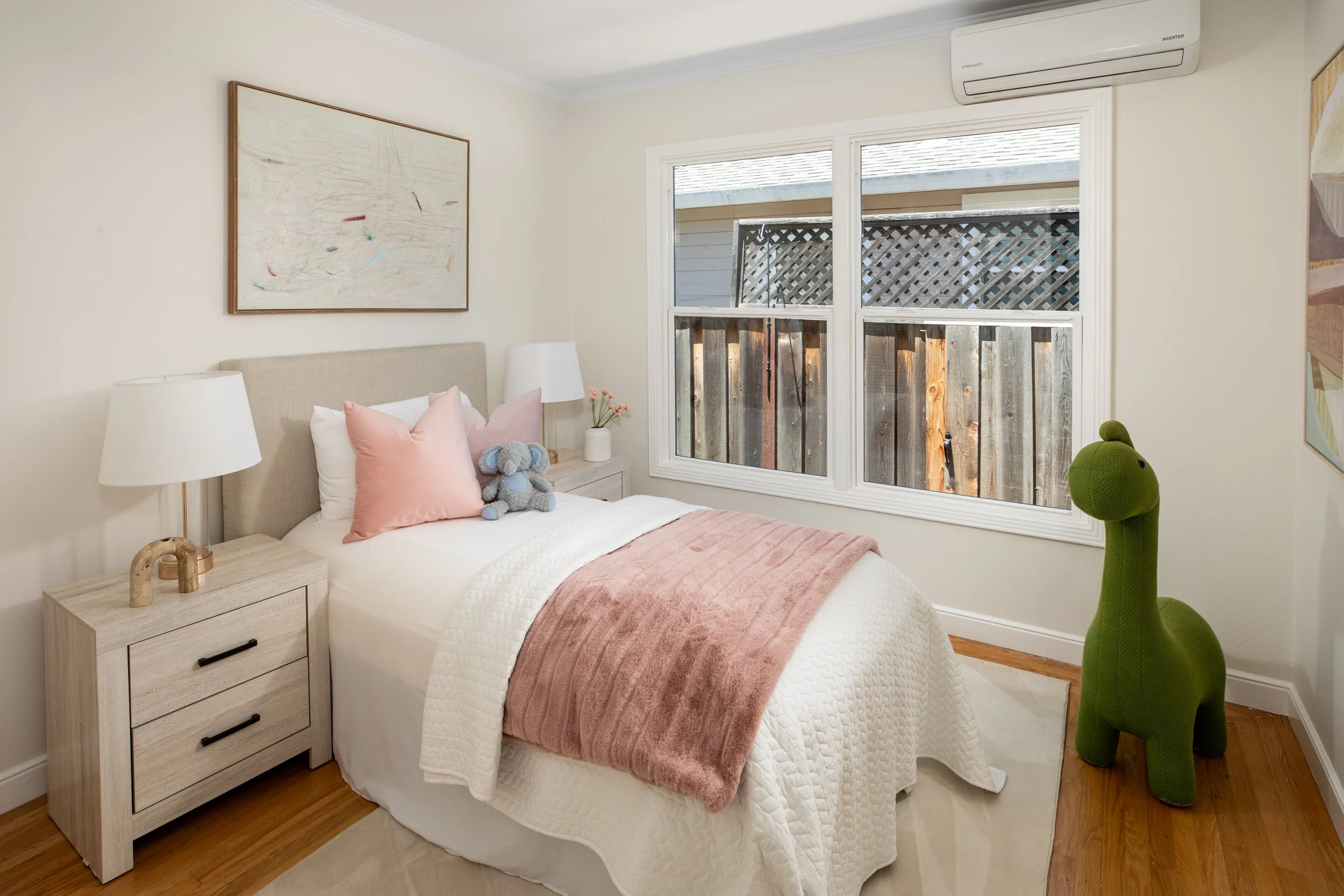 A cozy bedroom with a bed that has pink pillows and a plush gray elephant toy. The nightstand beside the bed holds a lamp and a small vase with pink flowers. A white window allows natural light, and a green stuffed dinosaur stands next to the window.