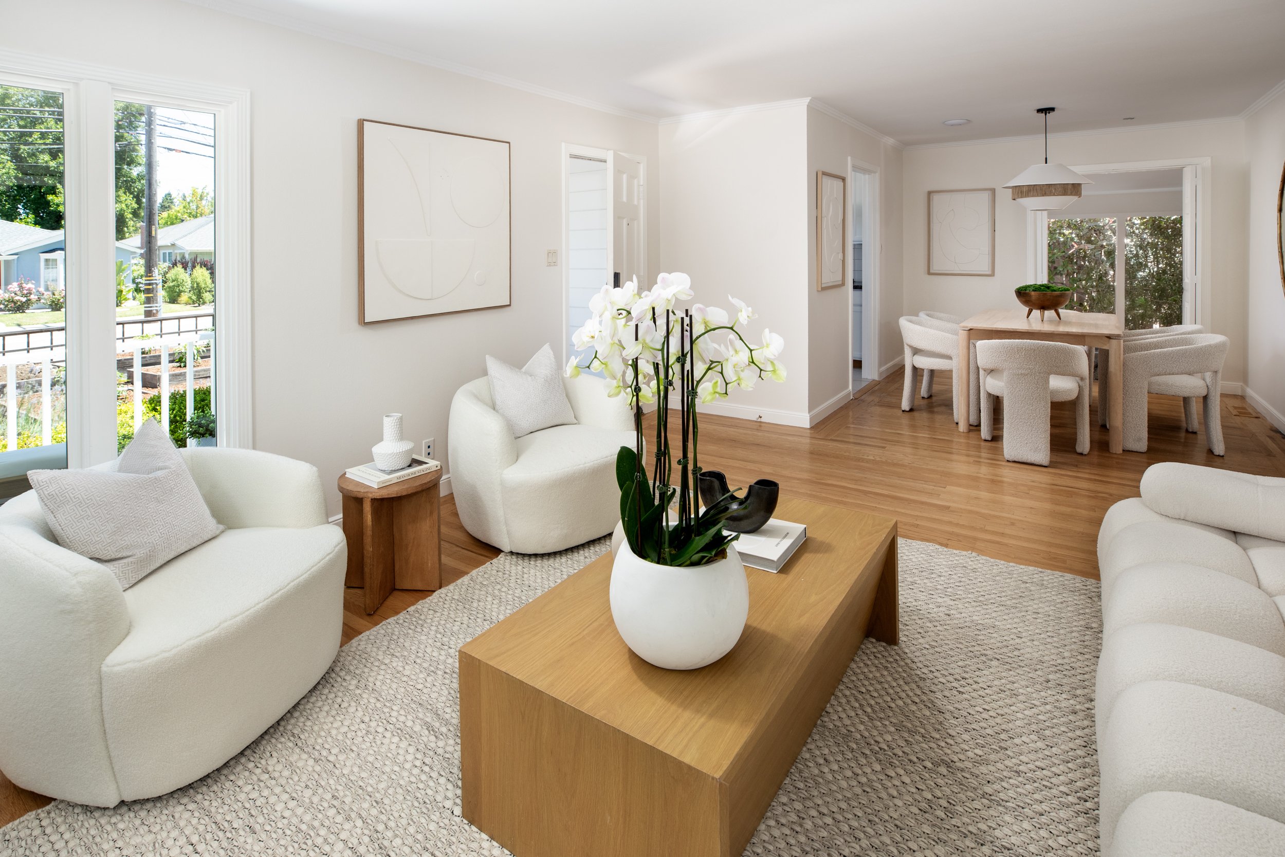 Bright, modern living room with white furniture, wooden flooring, a large window, and minimalist decor including wall art and a plant on the coffee table.