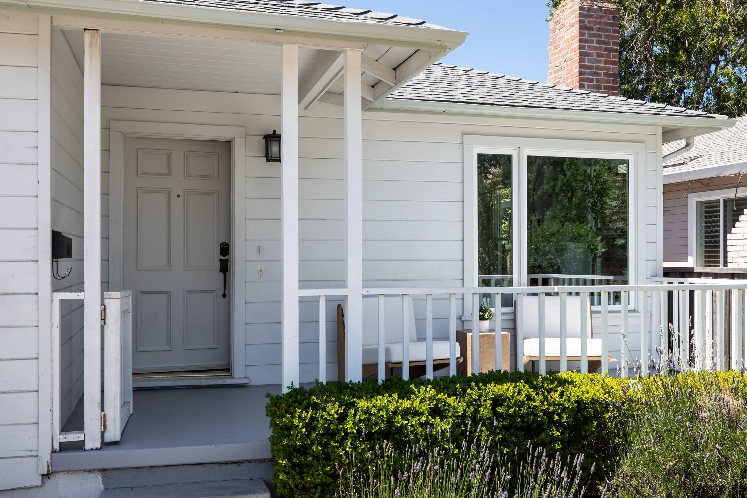 White house entrance with a porch, white railing, bushes, and large window.