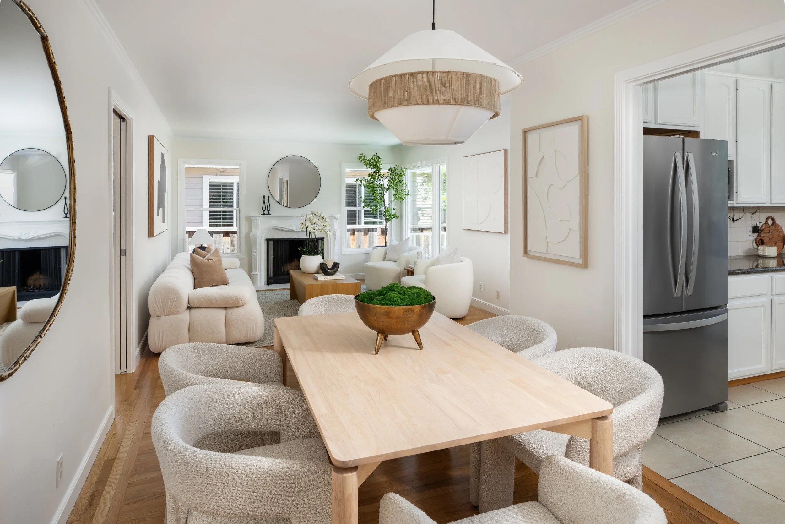 Interior view of a bright living and dining room with white walls, light wood flooring, and a wooden dining table with six textured cream-colored chairs. There is a white sofa and two white armchairs in front of a fireplace, which has a white ornate 