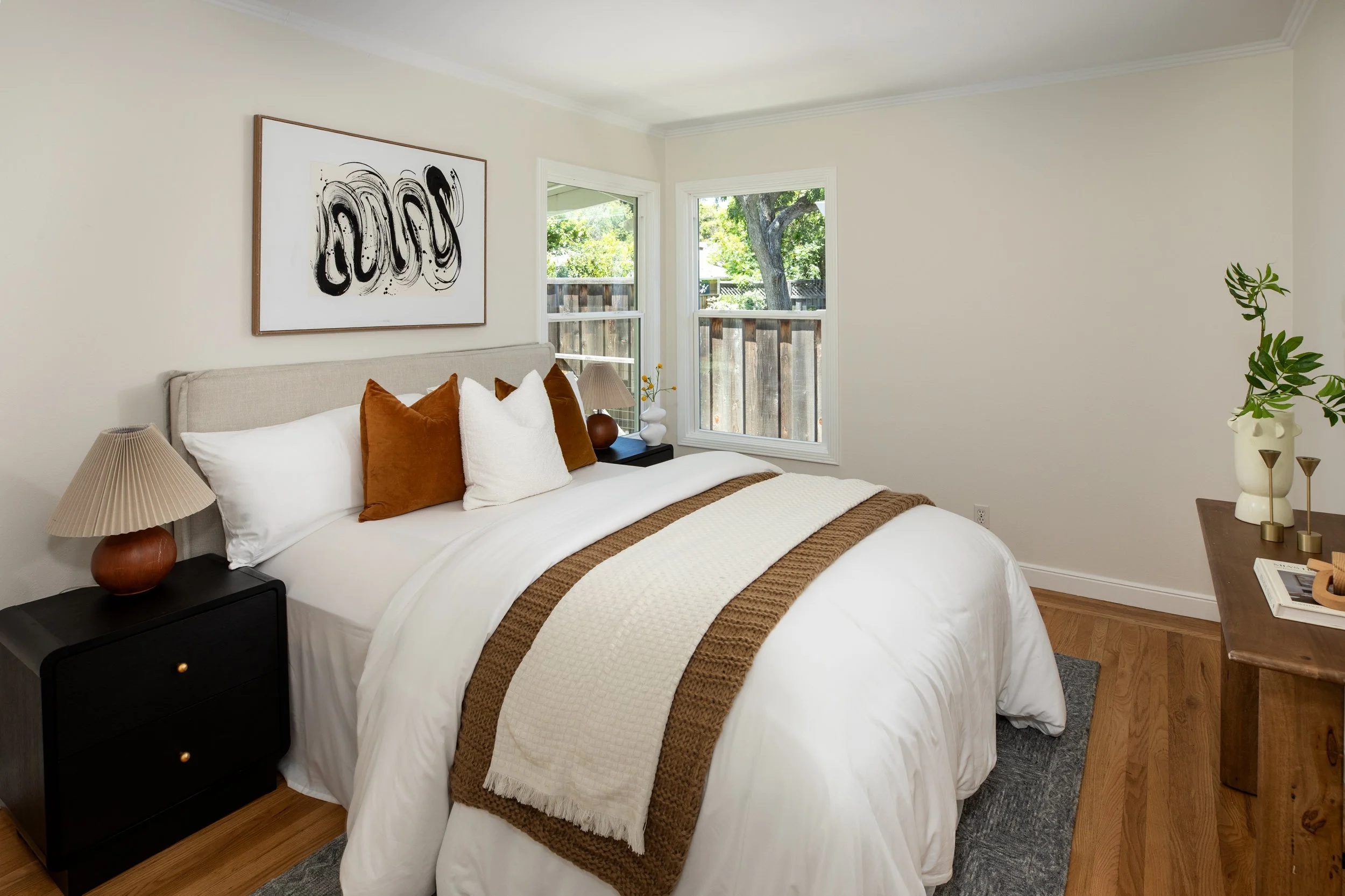 A bedroom with a bed, nightstand, lamps, and art on the wall, with windows showing trees outside.