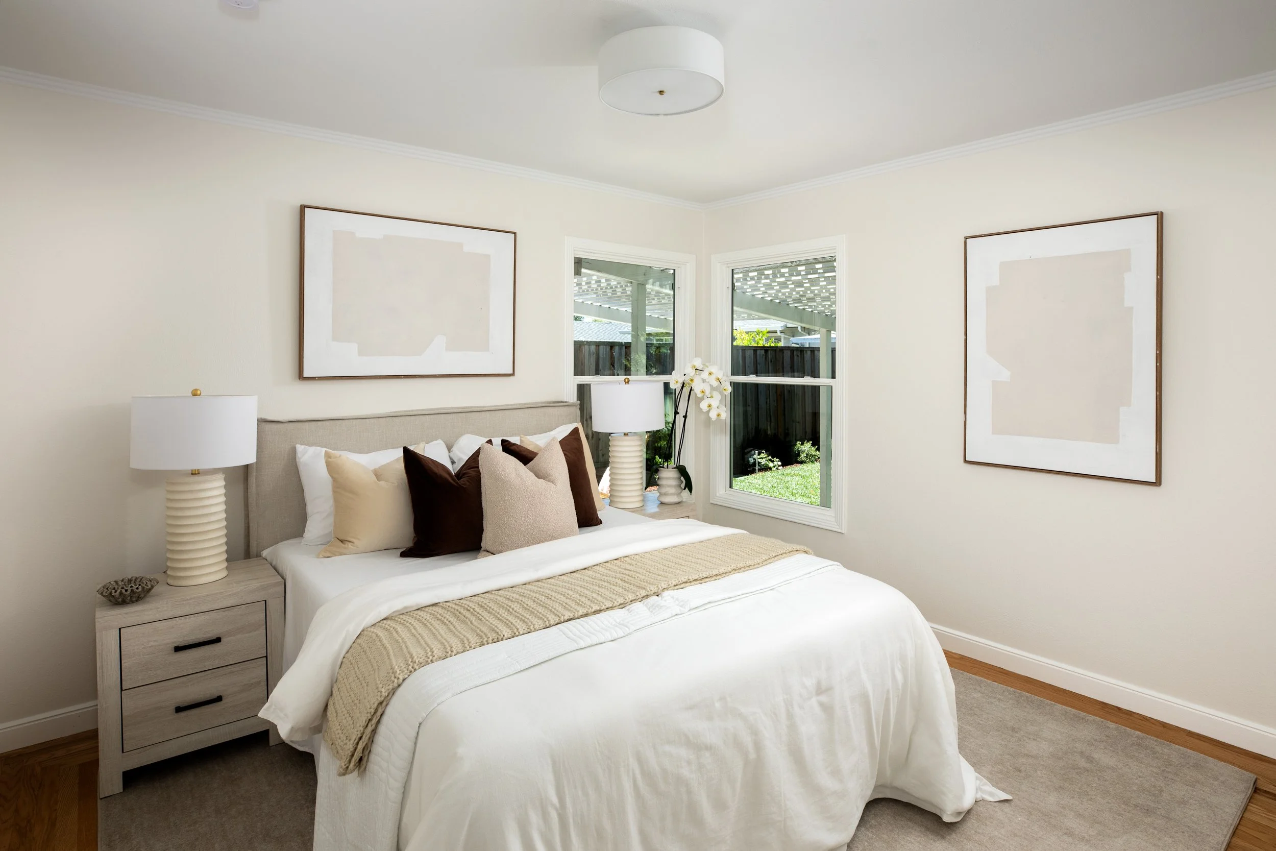 A neatly made bed with white bedding, multiple beige and brown pillows, and a beige throw blanket in a bedroom with cream-colored walls and two abstract framed artworks. There are two matching white lamps on wooden nightstands and two windows showing