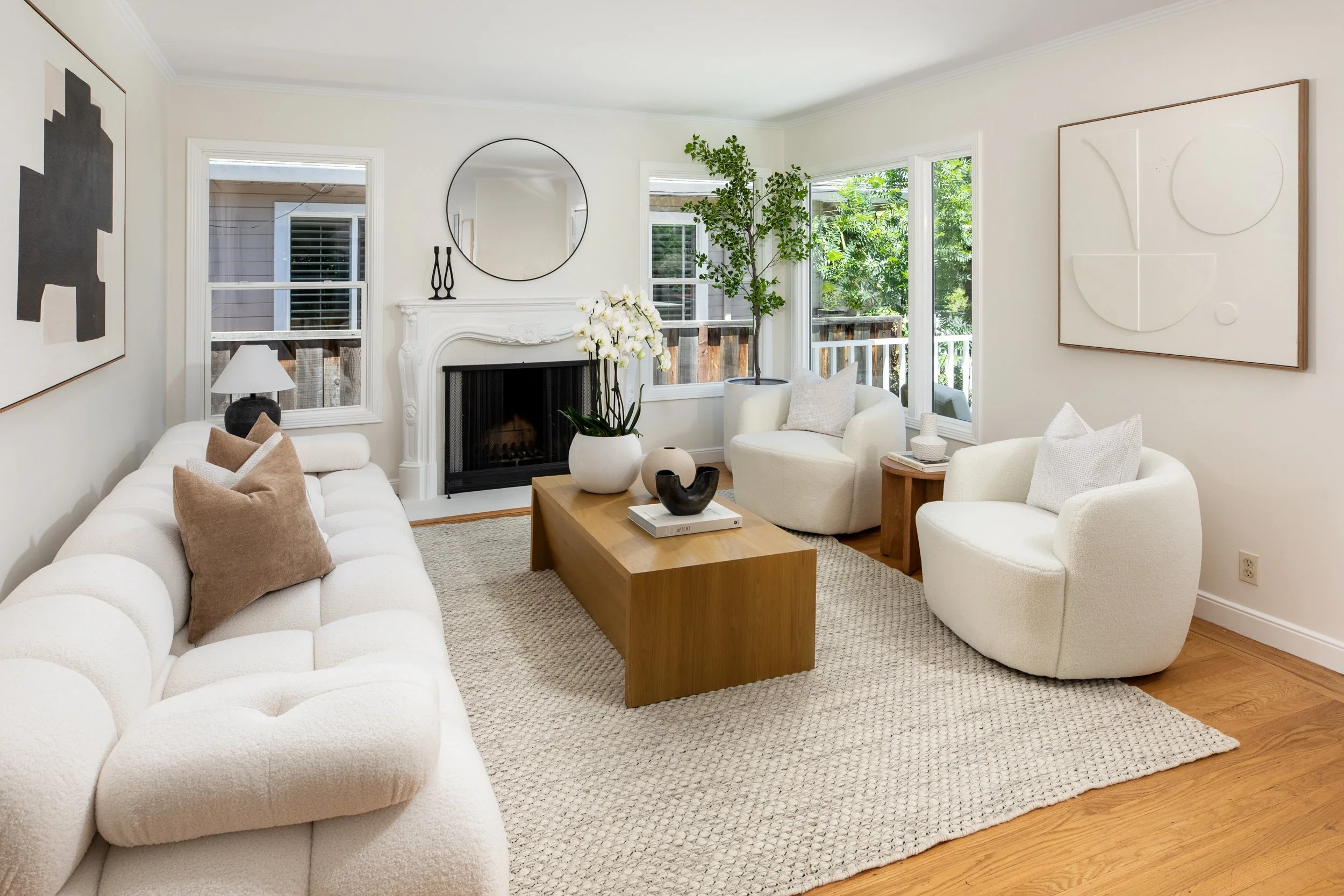 A cozy living room with a white couch, beige and white pillows, a wooden coffee table with decorative vases, a white armchair with a cushion, a fireplace with a round mirror above, green plants near large windows, and abstract wall art, all in a brig