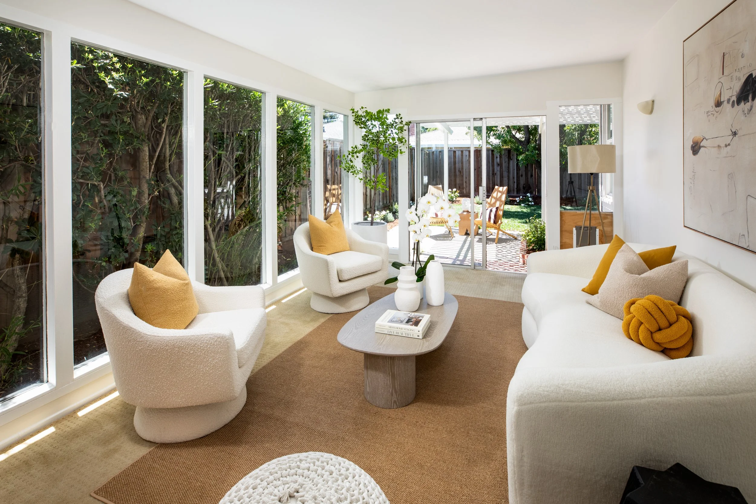 Living room with large windows, white sofas and armchairs with yellow pillows, a wooden coffee table with books and white vases, and a sliding glass door leading to an outdoor patio with chairs and a garden.