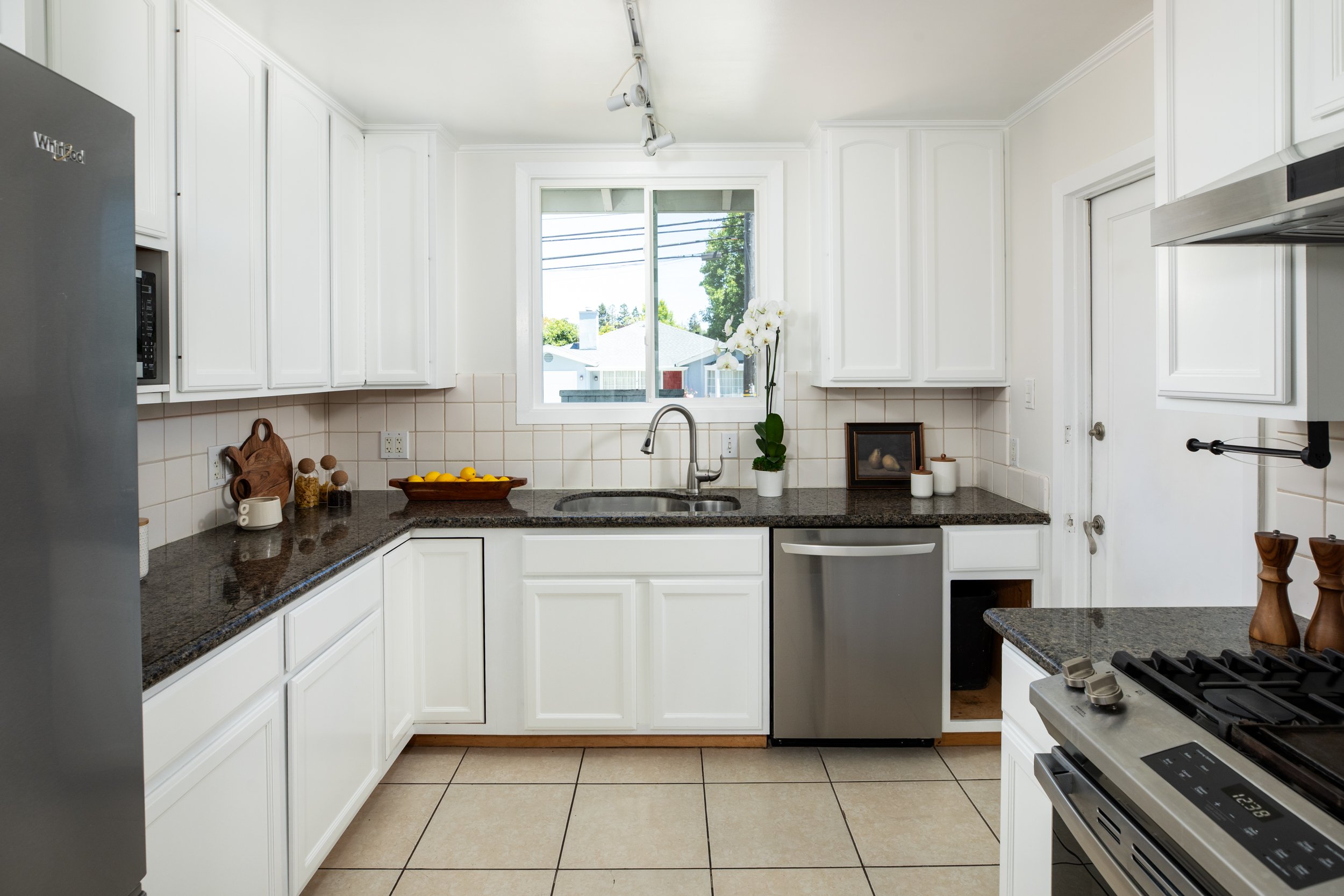 A clean white kitchen with white cabinets, a granite countertop, a stainless steel dishwasher, a window above the sink, a orchid plant, and some framed pictures.