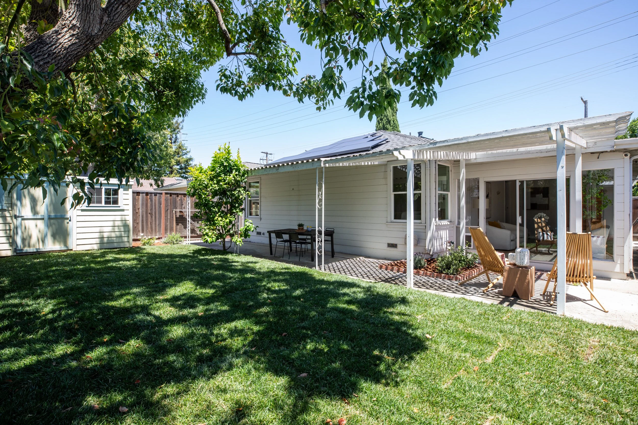 Backyard with a white house, patio furniture, and a grassy lawn shaded by large trees on a sunny day.