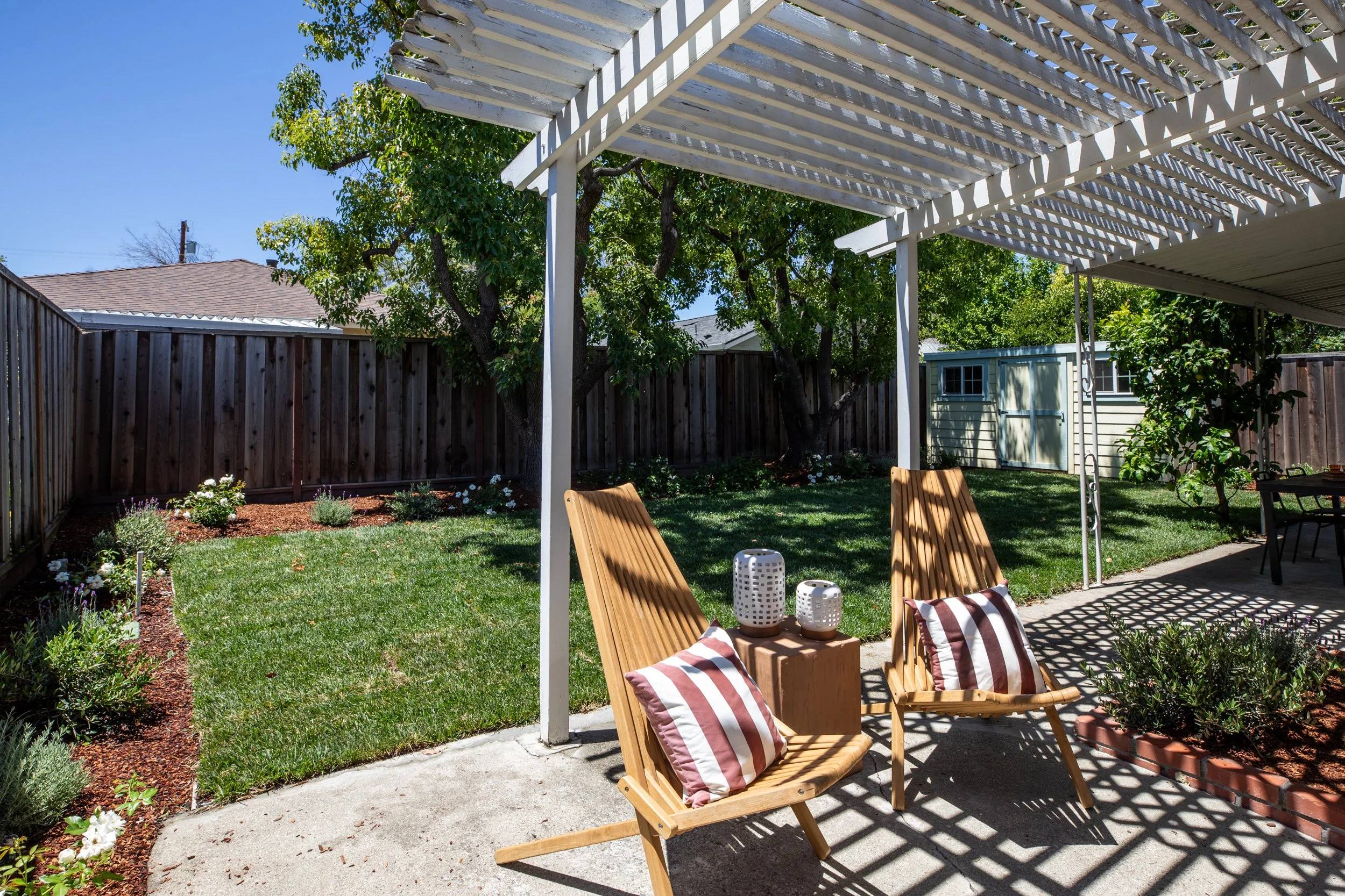Backyard patio with wooden chairs, striped pillows, small decorative lanterns, a pergola casting shadows, green grass, flower bed, trees, wooden fence, garden shed, clear blue sky.