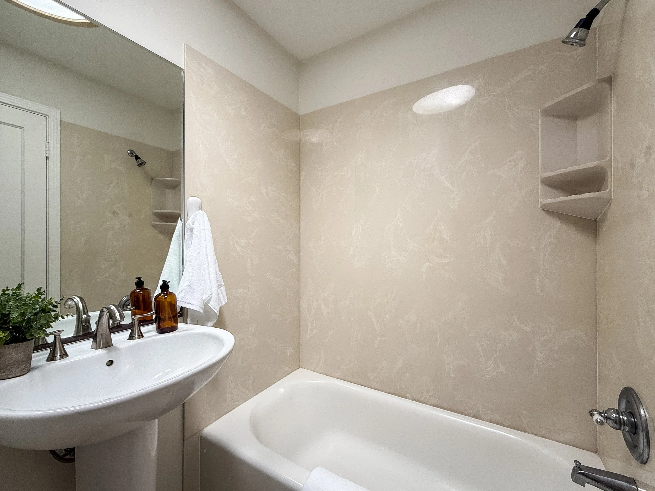 Bathroom with a bathtub, a pedestal sink with two amber bottles, a mirror, a towel hanging, beige marble wall, and shower fixtures.