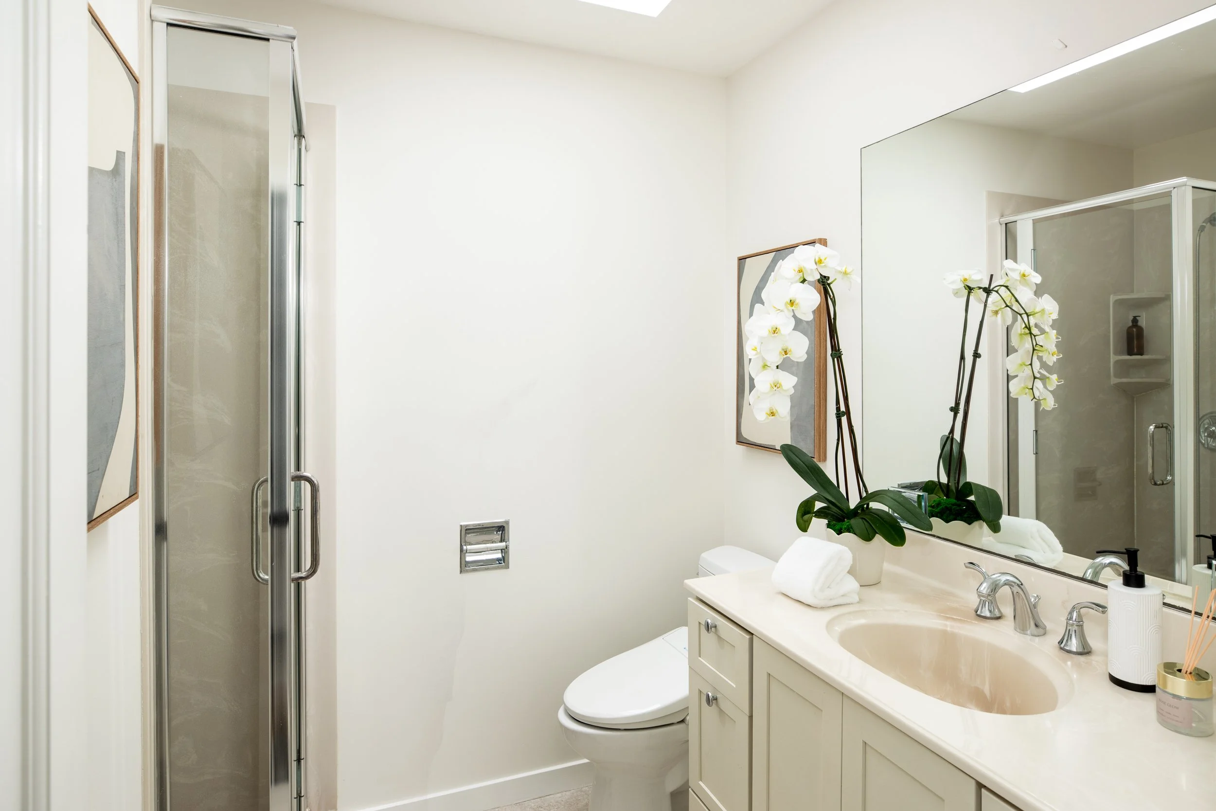 A bathroom with a shower stall on the left, a white toilet below a large mirror, and a cream-colored vanity with a sink, soap, a rolled towel, and decorative orchids in a pot.