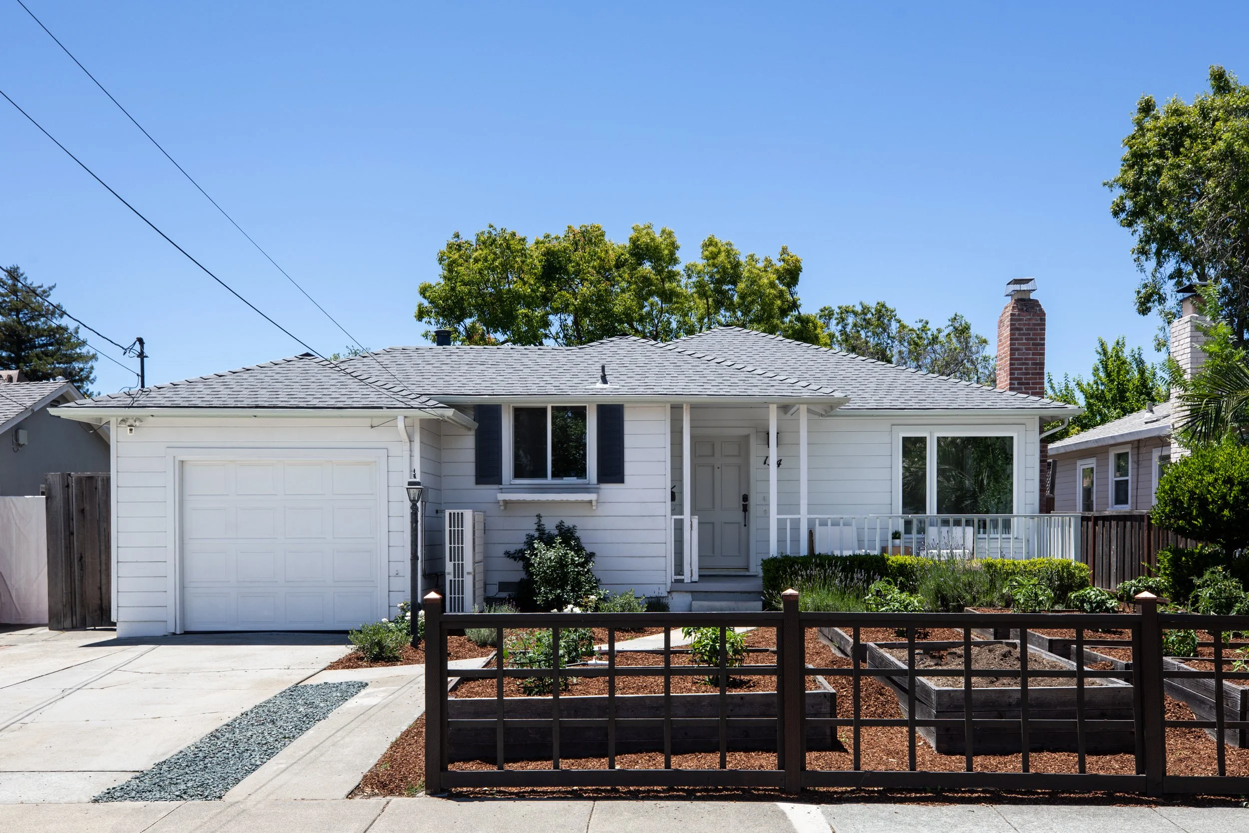 White house with attached garage and chimney, fenced garden with plants, dry mulch, sidewalk, and trees in background, under a clear blue sky.