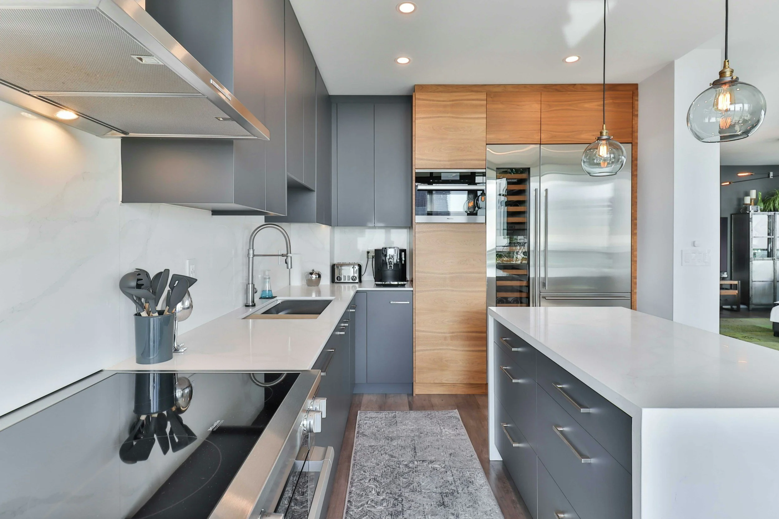 Modern kitchen with gray cabinets, white countertops, stainless steel appliances, wooden accents, hanging glass pendant lights, and a gray rug on dark hardwood floor.
