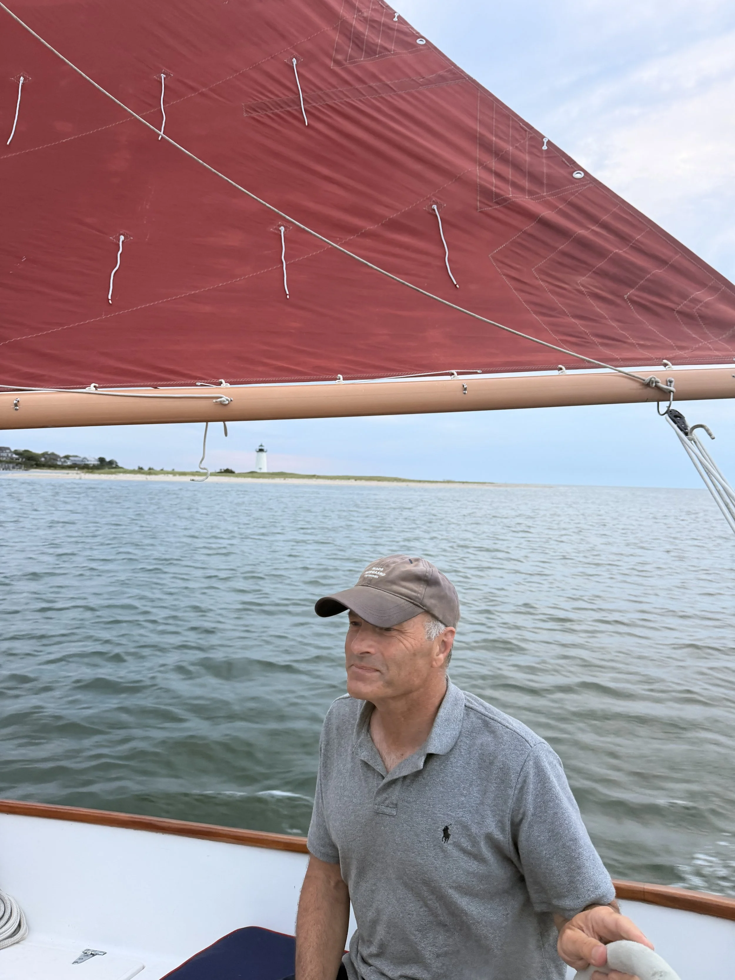 A man with gray hair and wearing a gray polo shirt is sitting on a boat, holding what appears to be a fish. In the background, there is a body of water with a lighthouse and land with trees and buildings in the distance. The sky is partly cloudy.