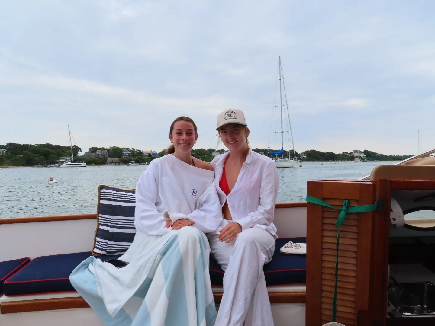Two smiling women sitting on a boat with water and sailboats in the background.