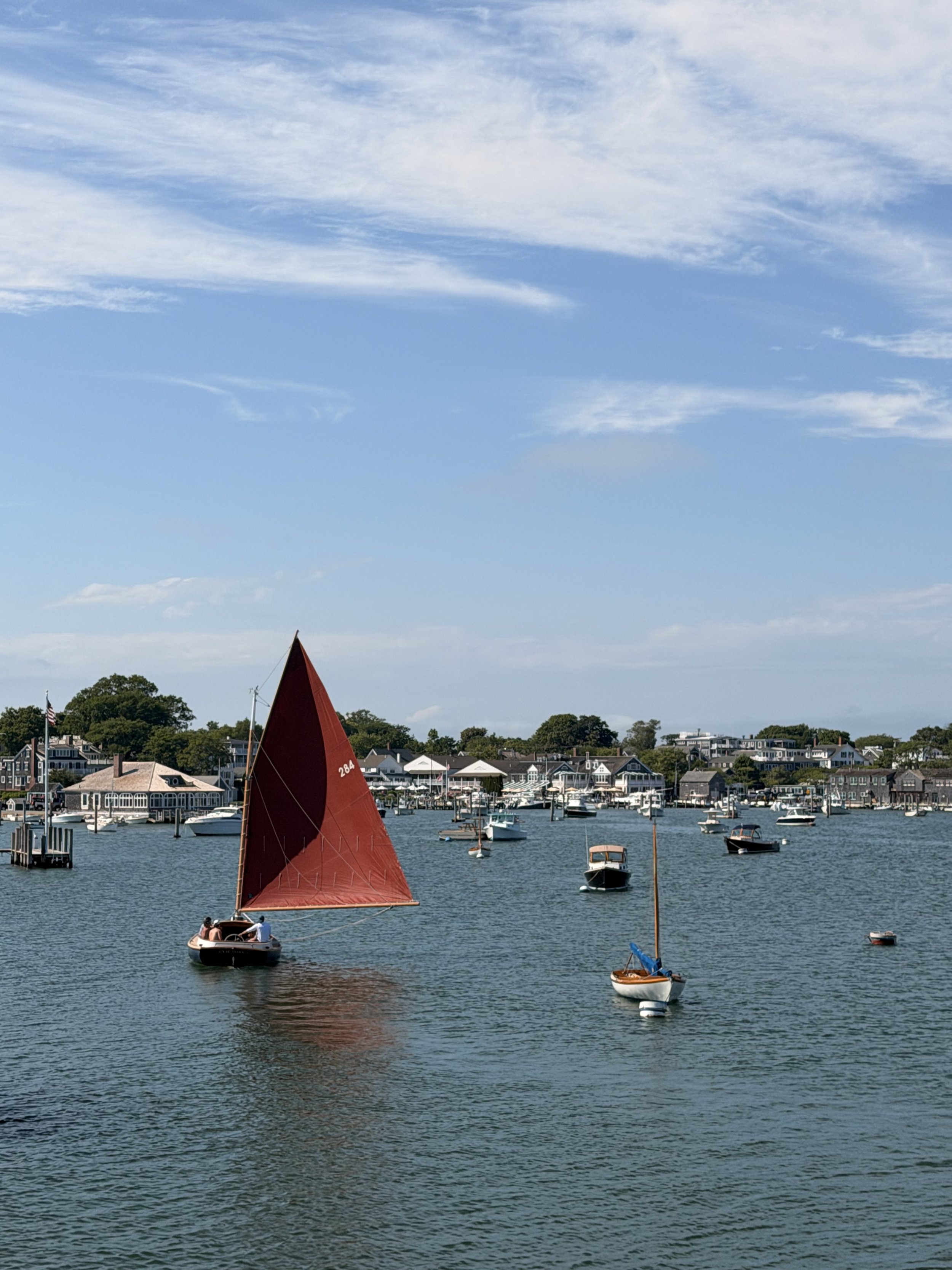 View of a harbor with several boats, including a small sailboat with a large red sail, and houses along the shoreline under a partly cloudy sky.