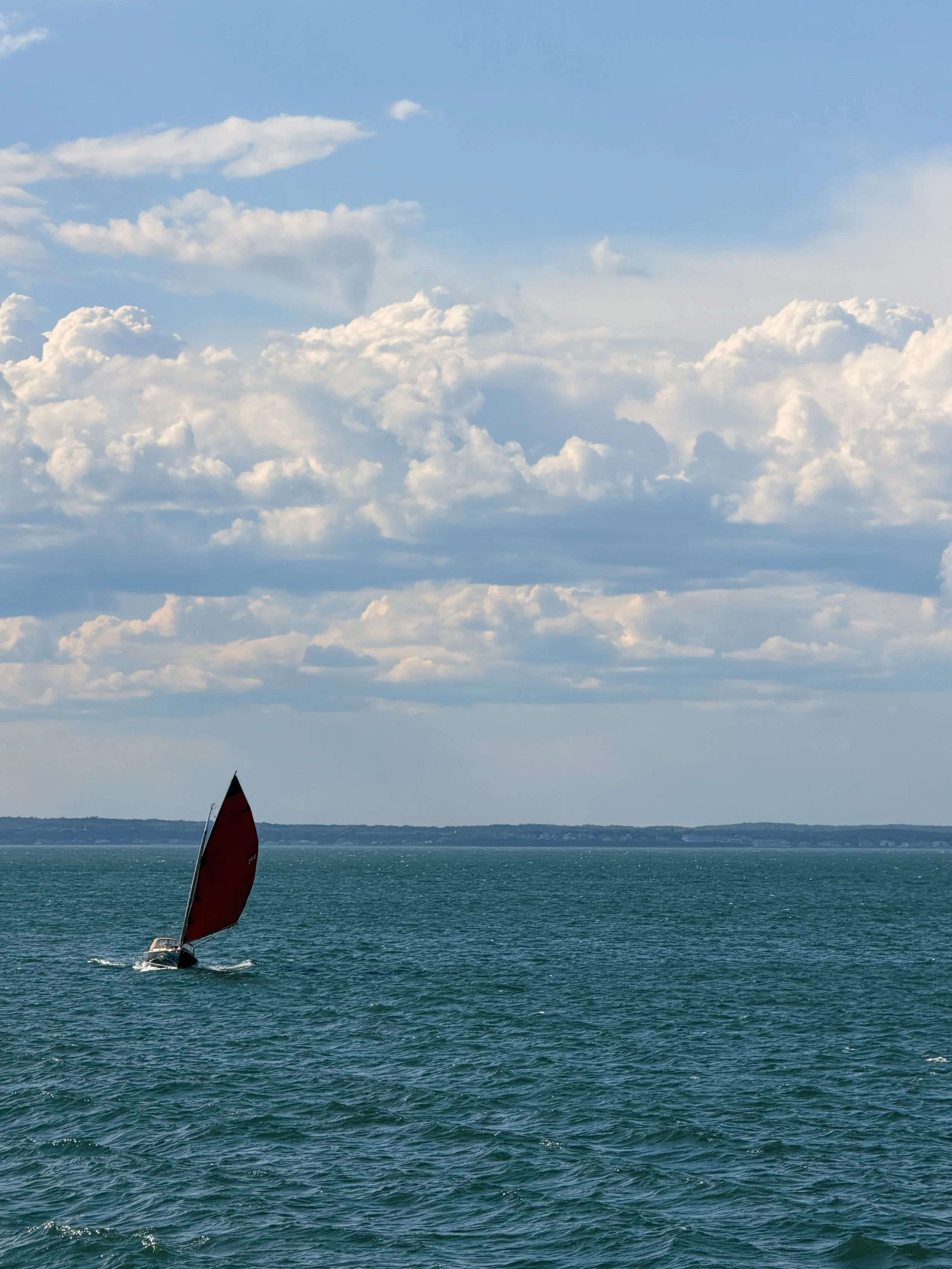 A sailboat with a red sail on the ocean with a hilly shoreline and partly cloudy sky in the background.