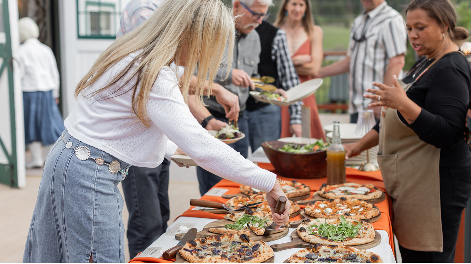 People serving and selecting pizza at a buffet-style outdoor gathering with salads and drinks.