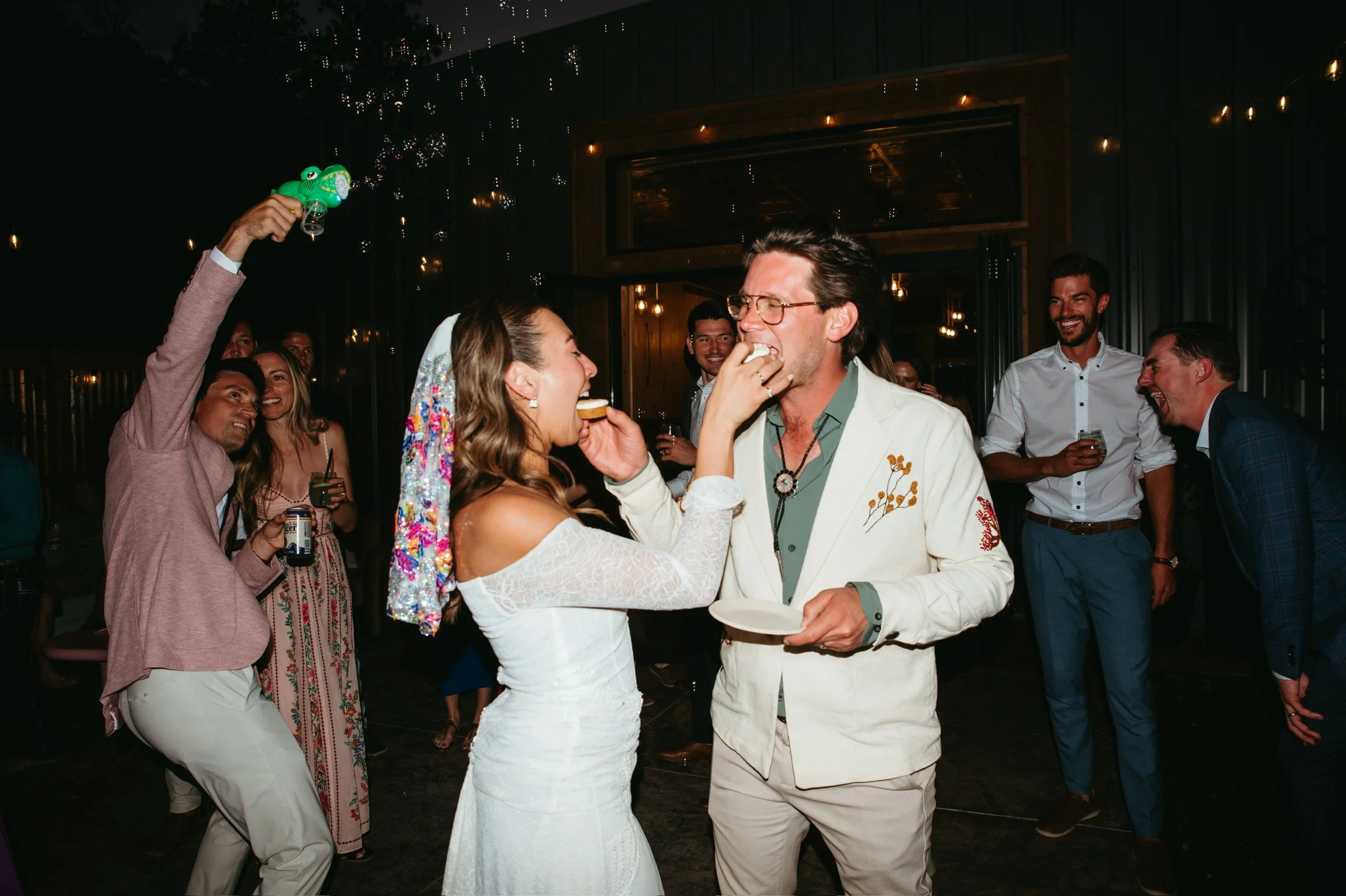 A bride and groom share a playful moment during their wedding reception, with the bride touching the groom's face as he smiles. The bride wears a white lace dress and a colorful veil, while the groom is dressed in a white suit with floral embroidery. Around them, guests are laughing and enjoying the celebration, some holding drinks. The setting features dim lighting and warm overhead lights.