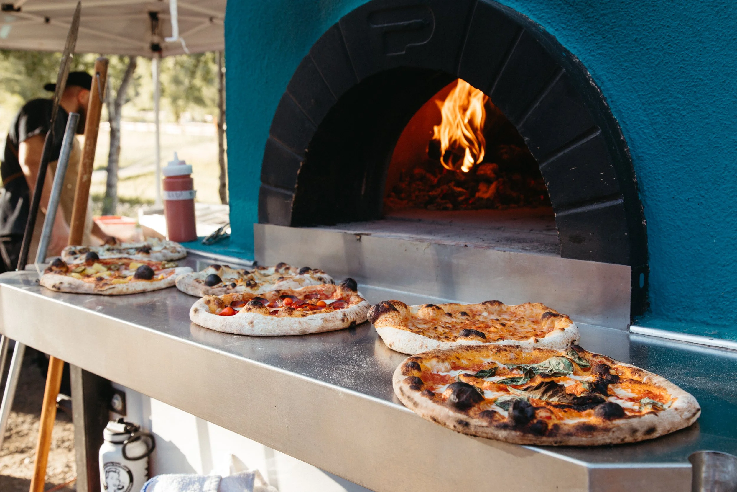 A pizza oven with a visible fire inside. Several pizzas with various toppings are placed on a metal counter in front of the oven, ready to be baked.